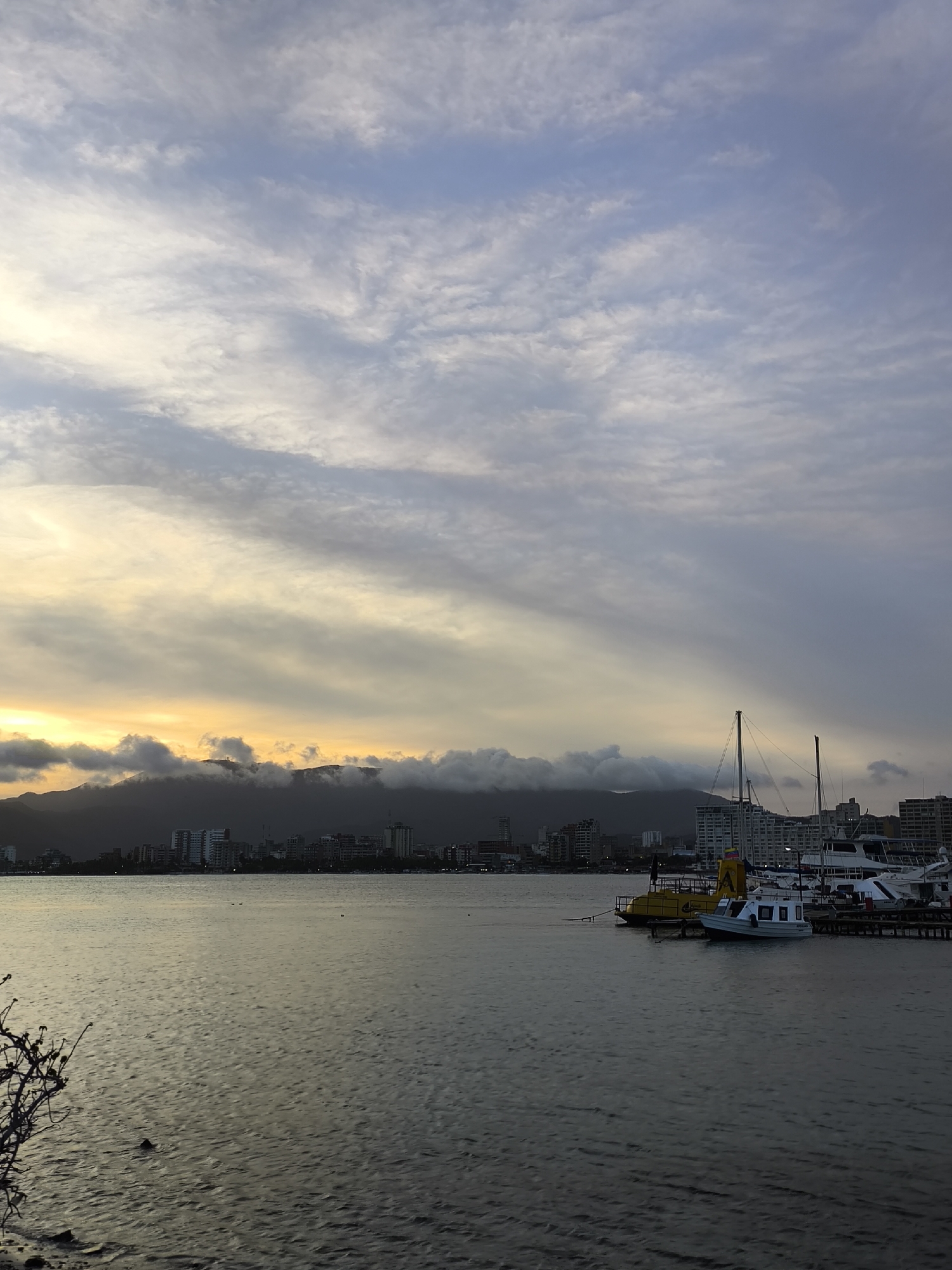 The image depicts a waterfront scene in Porlamar, Venezuela, at dusk or dawn. The sky is filled with layered clouds, ranging from light blue and white at the top to darker gray and yellow hues closer to the horizon. In the distance, a mountain range is partially obscured by low-hanging clouds.
A city skyline with numerous buildings is visible along the shoreline. In the foreground, a body of water reflects the colors of the sky. Several boats, including a yellow submarine-like vessel, are docked at a pier to the right. Bare branches are visible in the lower left corner, suggesting vegetation near the viewer's location.