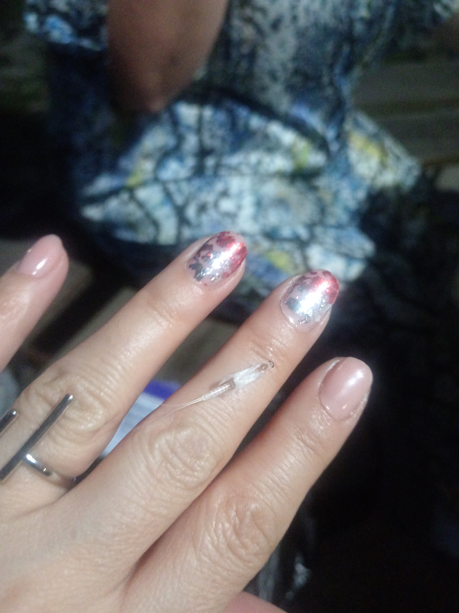 The image shows a close-up view of a person's hand, featuring manicured nails with red and silver glitter polish. A thin, transparent tube or filament is attached to the middle finger, extending towards the viewer. The background is blurred, but appears to be a patterned fabric or surface. The image does not contain any information that can be used to determine its location in the city of Belogorsk, Russia.