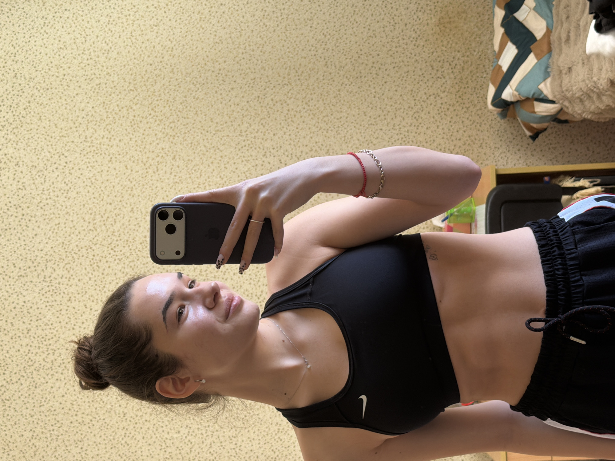 A young woman is taking a selfie in an indoor setting. She is wearing a black Nike sports bra and black shorts with a white stripe and drawstring. She is holding a black smartphone with multiple lenses, displaying it in front of her face. She has bracelets on her left wrist and a ring on her left hand. Her hair is pulled back in a bun, and she has a necklace on. The background includes a yellow patterned wall and part of a bed with pillows and a folded blanket. There is also a glimpse of a dark piece of furniture. The location cannot be determined.