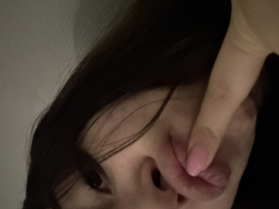 A selfie shows a person with dark hair, fair skin, and an eye visible. A hand with a pink nail touches their lips, partially obscuring their face. The angle of the photo is tilted, with the person's head positioned in the upper left corner of the frame. The background appears to be a light wall.