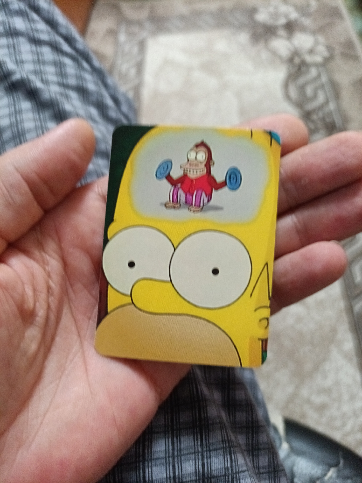 A close-up, overhead shot captures a hand holding a square trading card featuring a cartoon character. The card depicts the upper portion of a yellow, bald character's head, with large white eyes and a wide smile revealing teeth. Inside a thought bubble above the character's head, a small, brown, ape-like creature with bulging eyes is shown lifting blue dumbbells. The background consists of a plaid fabric, likely clothing, and a blurred, textured surface. The image does not contain any information about the city of Nyagan, Russia, or any other geographical location.