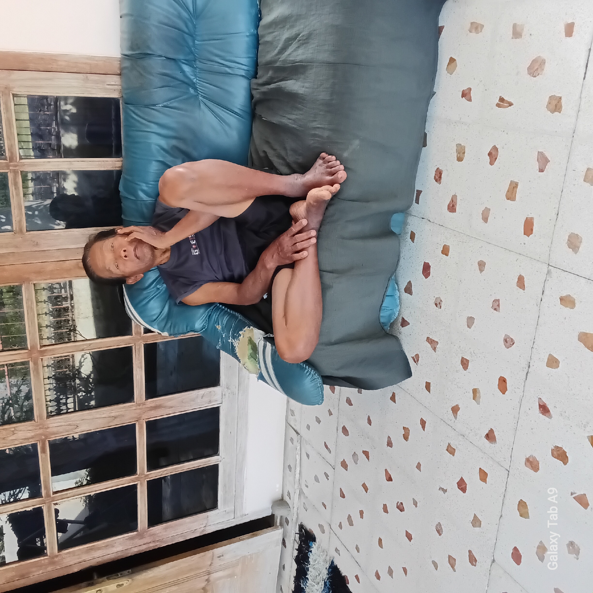 An elderly man is sitting on a turquoise sofa with a grey blanket draped over the front. He is wearing a dark grey t-shirt and black shorts. The man is resting his left hand on his cheek and his legs are crossed. The sofa is situated on a tiled patio with a speckled white and brown pattern. In the background, a wooden door with multiple glass panes is visible, reflecting the outside environment which includes greenery and what appears to be a fence. The scene is outdoors, likely in a residential area in Sambirejo, Indonesia. The overall lighting suggests it is daytime.