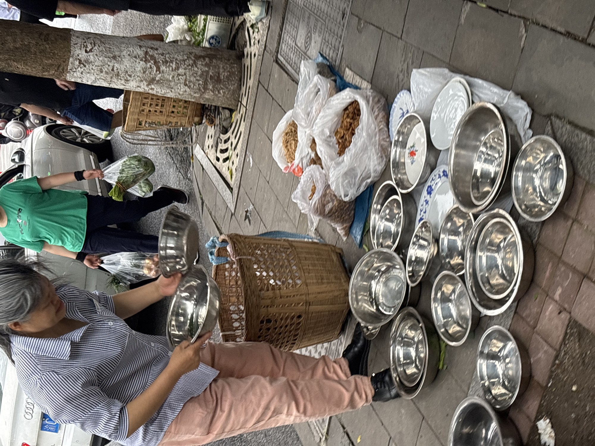 On a sidewalk in Jiangjin, China, an Asian woman with gray hair, wearing a striped blue and white shirt and pink pants, sits on the ground, holding two stainless steel bowls. She appears to be examining or preparing them. Around her are numerous other stainless steel bowls of varying sizes, arranged on the ground. Next to the bowls are bags of dried food items and a woven basket. A man in a green shirt and dark pants stands nearby, holding a bag of leafy greens. A tree is to the left of the woman, and a parked car is partially visible behind her. Other people are visible in the background. The sidewalk is made of paving stones. A metal grate is visible on the sidewalk near the tree.