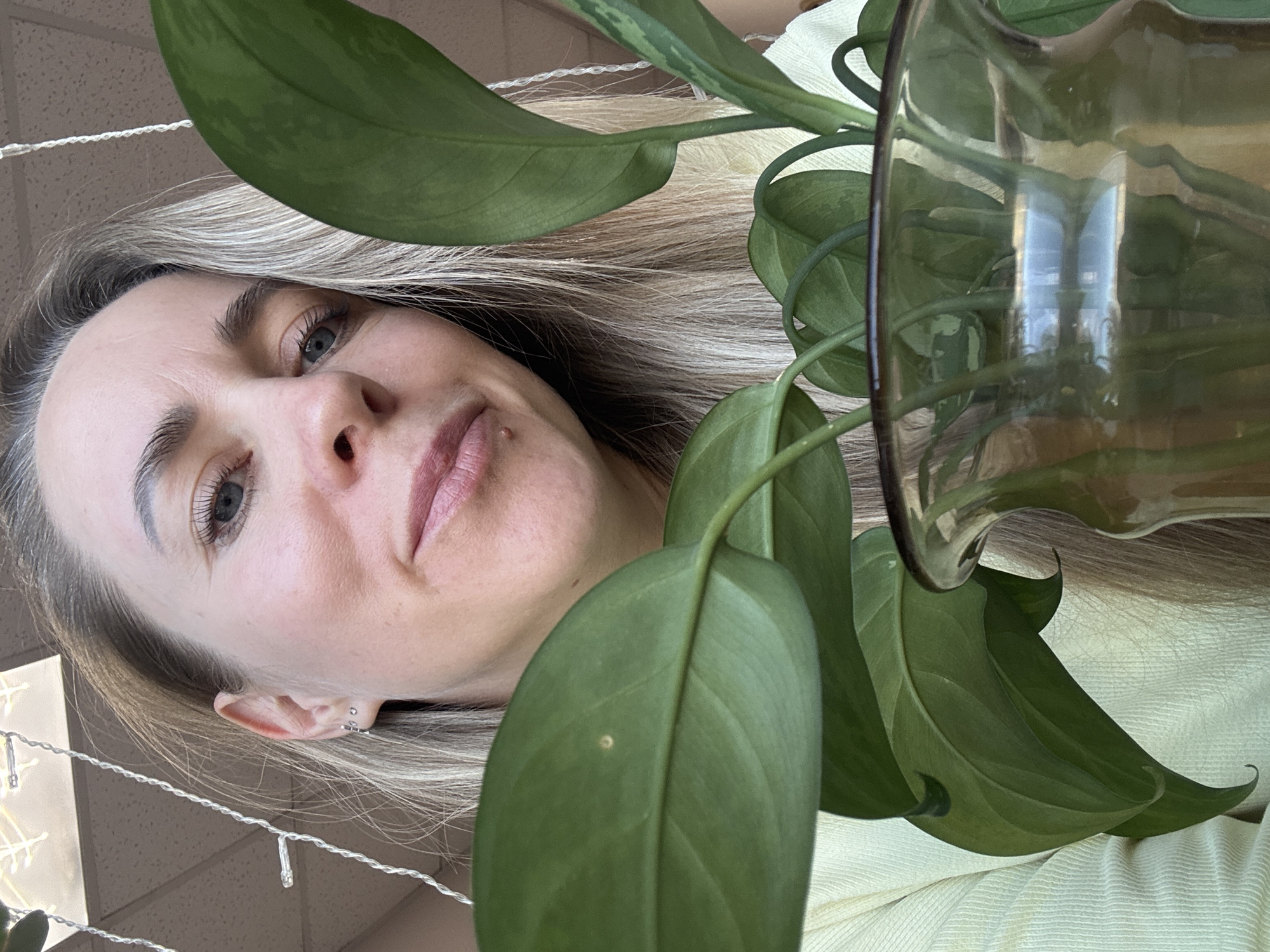 A woman with blonde hair and light skin is holding a plant in a glass vase. The woman is looking at the camera with a slight smile. She has multiple piercings on her left ear. The plant has large green leaves. There are decorative lights hanging in the background. The specific location in the city of Chaikovsky, Russia cannot be determined from this image.