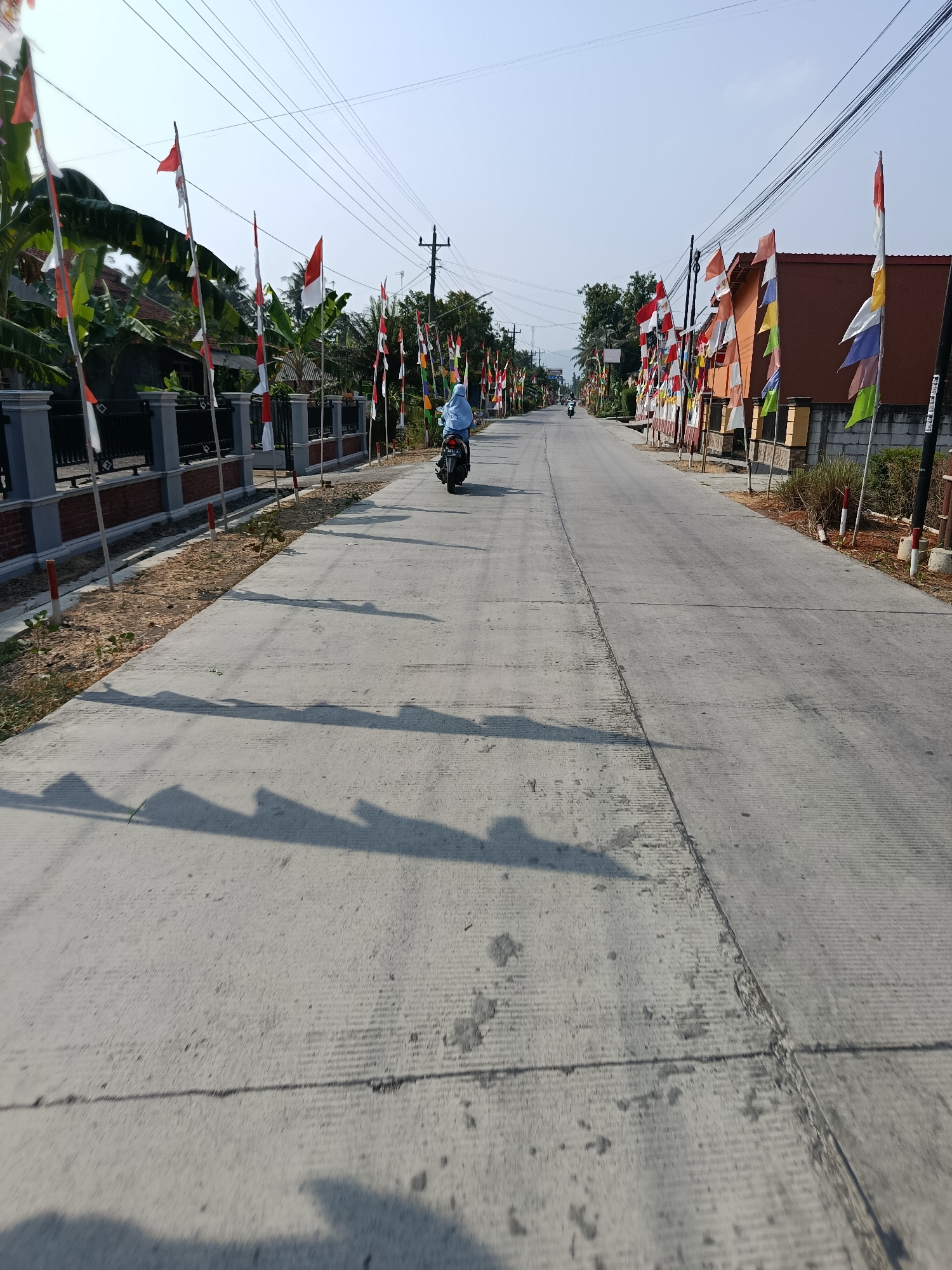 In Sarwadadi, Indonesia, a paved road stretches into the distance under a clear, bright sky. The road is lined with numerous Indonesian flags, predominantly red and white, decorating the sides of the street and the fronts of buildings. A person is riding a dark-colored motorcycle down the road, wearing a light blue headscarf and clothing. The environment appears to be a semi-rural or suburban area, with houses and trees visible on either side of the road. The sun casts long shadows from the flags and possibly other unseen objects onto the concrete road surface. Power lines are visible strung across the sky. The overall scene suggests a festive or patriotic occasion, given the prevalence of the national flags.