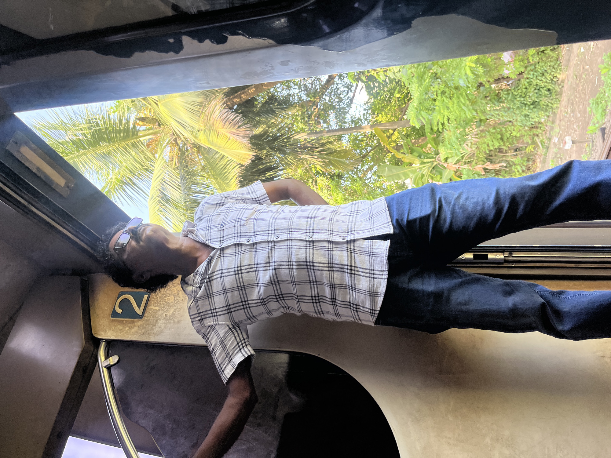 A man stands in a train carriage, looking out of an open doorway at lush greenery and palm trees, indicative of Aluthgama, Sri Lanka. The man, wearing a checkered shirt and dark jeans, has his left arm resting on the interior of the carriage and his right hand behind his back. He is looking upwards and to the right, with sunglasses on. A prominent number '2' is visible on the wall behind him, likely indicating a seat or carriage number. The lighting suggests it is daytime, with sunlight filtering through the vegetation. The scene captures a moment of observation from within the moving train.