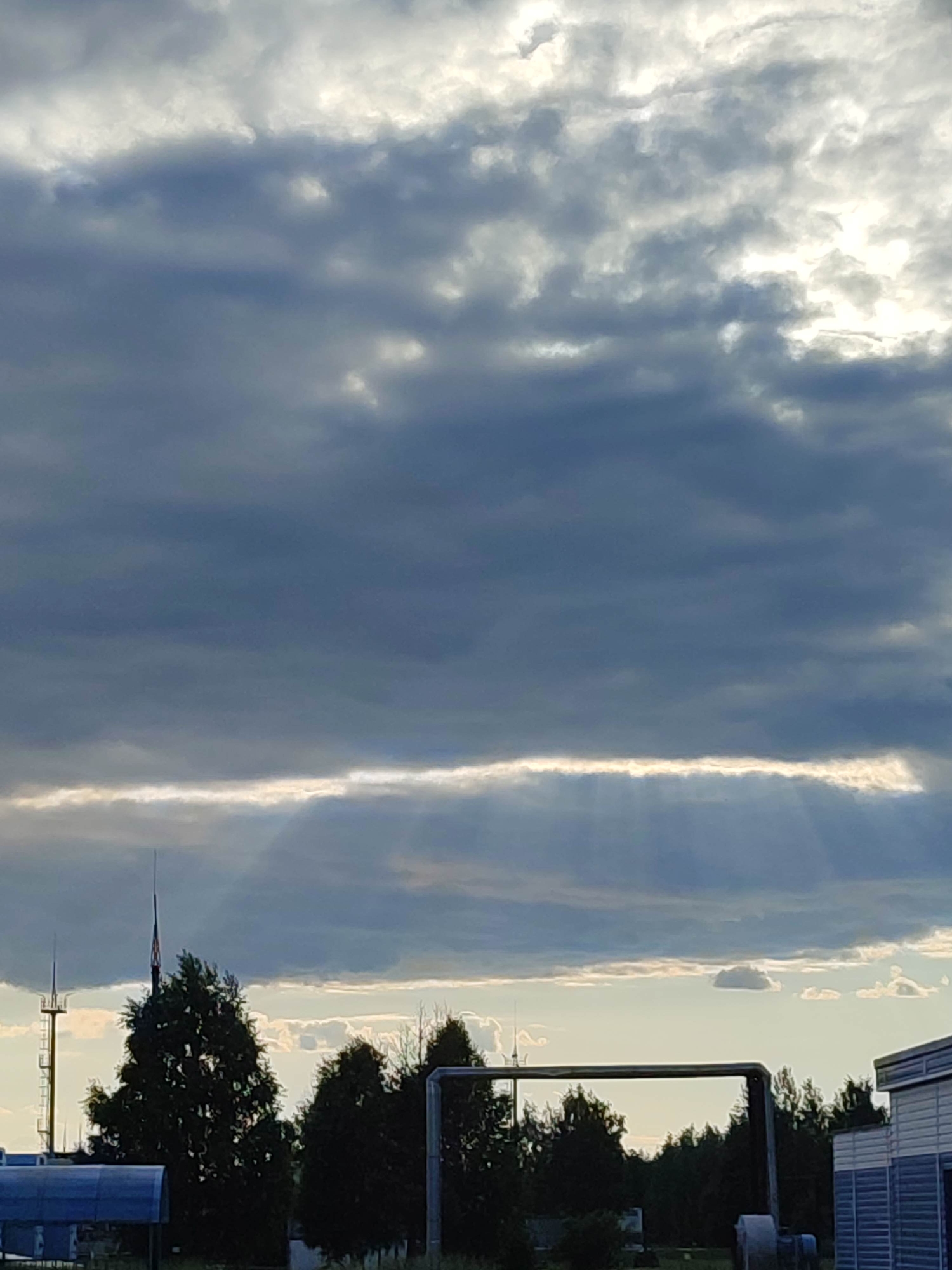 The image captures an outdoor scene under a heavily clouded sky, depicting the interplay of sunlight and cloud cover over an industrial or utility area. The upper two-thirds of the frame are dominated by dense, dark grey cumulonimbus clouds, through which patches of bright white light are visible, indicating the sun's position. A prominent, continuous horizontal band of brilliant white light stretches across the mid-section of the sky, where the cloud cover is thinner. Below this illuminated band, distinct crepuscular rays (sunbeams) are seen fanning downwards through the lower cloud layers. The horizon line features a dense row of dark green trees, silhouetted against a lighter, pale blue-grey sky.
In the foreground and midground, several man-made structures are visible. On the far left, a tall, slender metal mast or antenna with a small platform is present. To its right, a taller, dark, spire-like structure, possibly a lightning rod or flagpole, rises above the treeline, exhibiting faint reddish-orange details near its apex. A portion of a building with a blue, curved roof is visible in the lower-left corner. Towards the center-right, a large, dark, pipe-like structure forms a horizontal bar across the frame, with other piping elements partially visible on the ground level. On the far right, a rectangular building with horizontal blue and white siding is partially visible. The overall environment suggests an industrial or infrastructure-heavy setting on the outskirts of a populated area, in the city of Unknown, Unknown. No people are visible in the image.