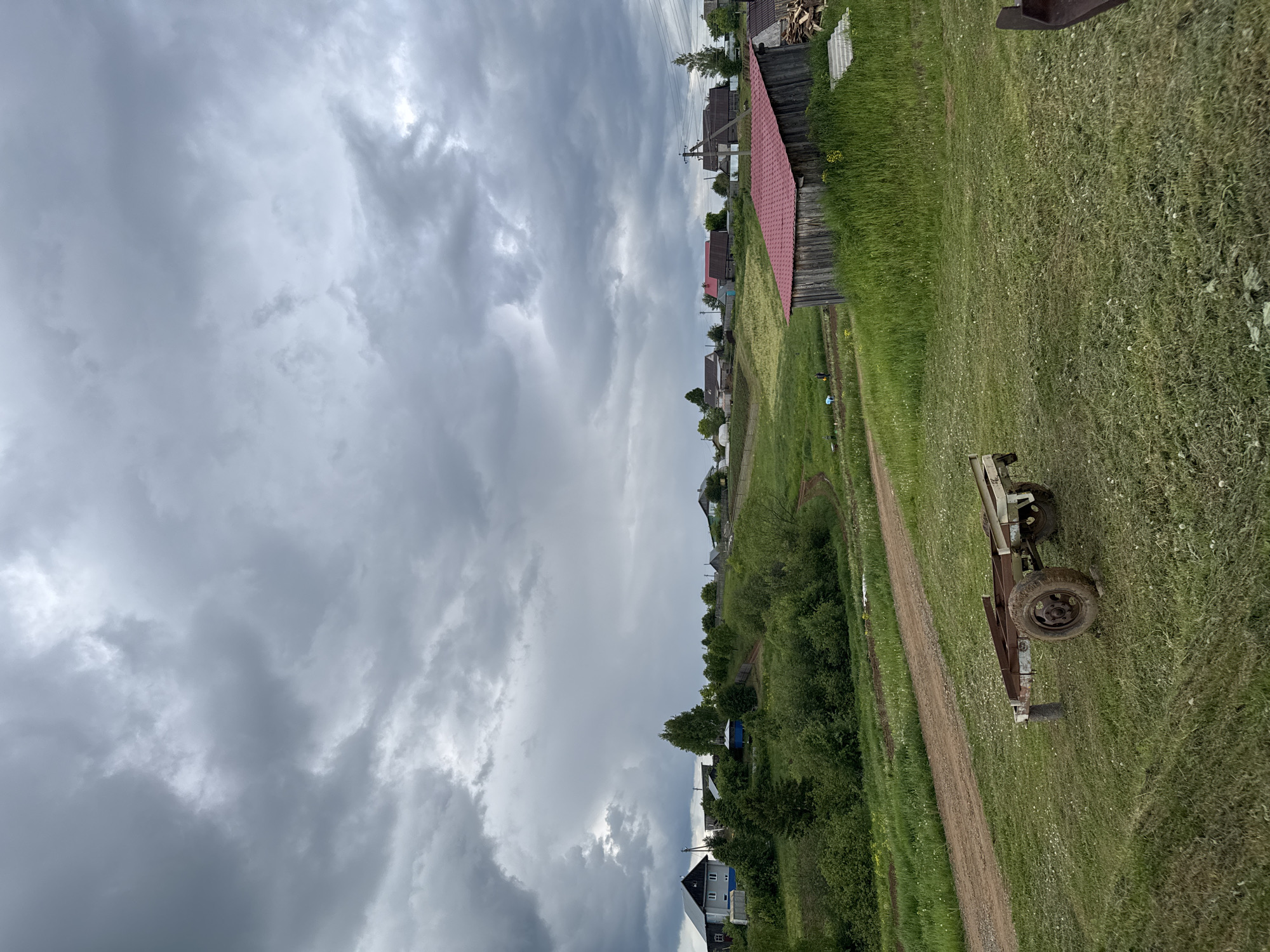 This landscape image captures a quiet, rural village scene under a dramatically overcast sky. A winding dirt path extends from the foreground into the middle distance, separating expanses of lush green grass and wild vegetation.
In the immediate foreground on the right, an old, rusty, two-wheeled trailer or cart is parked in the grass next to the path. Its weathered metal frame and robust wheel suggest functionality within an agricultural or rural context.
Beyond the path, a collection of houses, indicative of a small village or hamlet, stretches across the mid-ground and into the background. These dwellings feature various roof colors, including distinct reddish-brown and darker tones, nestled among scattered trees and bushes. Utility poles with overhead lines are visible, providing infrastructure to the remote settlement. Fences and what appear to be piles of firewood next to some houses add to the rustic character.
The sky dominates the upper half of the image, filled with heavy, dark and light grey clouds. This dense cloud cover creates a diffused light, suggesting an overcast day, possibly indicating an approaching storm or simply a perpetually gloomy weather condition. The absence of direct sunlight makes it difficult to pinpoint the exact time of day, but the overall mood is rather subdued and tranquil. No people or specific activities are visible, contributing to the serene and untouched feel of the scene. The setting aligns with a typical rural landscape found in locations like Бородулино, Russia. There is no discernible text within the image.