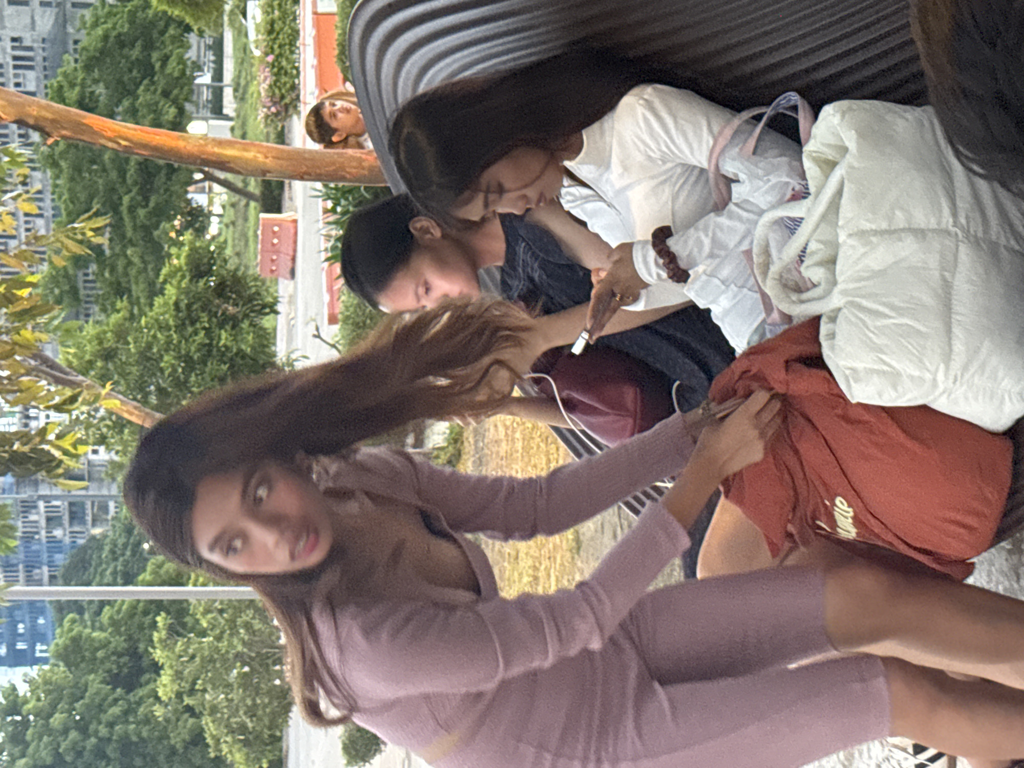 The image shows three individuals seated on a curved metal bench. The person closest to the camera is wearing a mauve-colored sweater dress and is facing the camera, with long brown hair tossed towards the left. Next to this person, two individuals are seated. One is wearing a white long-sleeved shirt, and the other is wearing a navy shirt. There is a white puffy vest, a maroon-colored tote bag, and another shopping bag on the bench. Outside the bench, there is a tree. In the distance, buildings and trees are visible. The location is Davao City, Philippines.