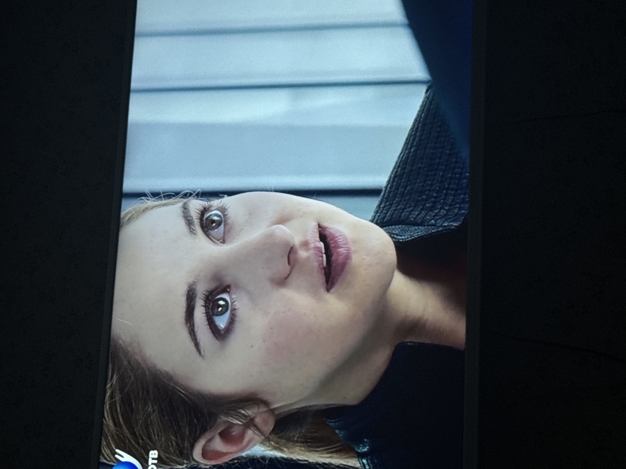 Close-up image of a woman's face, likely on a TV screen, revealing detailed facial features like brown eyes, freckles, and slightly parted lips. The background appears to show a blurred image of horizontal lines and possibly the interior of a vehicle due to the fabric texture near her neck. There are no discernible indicators of the city of Mytishchi, Russia.