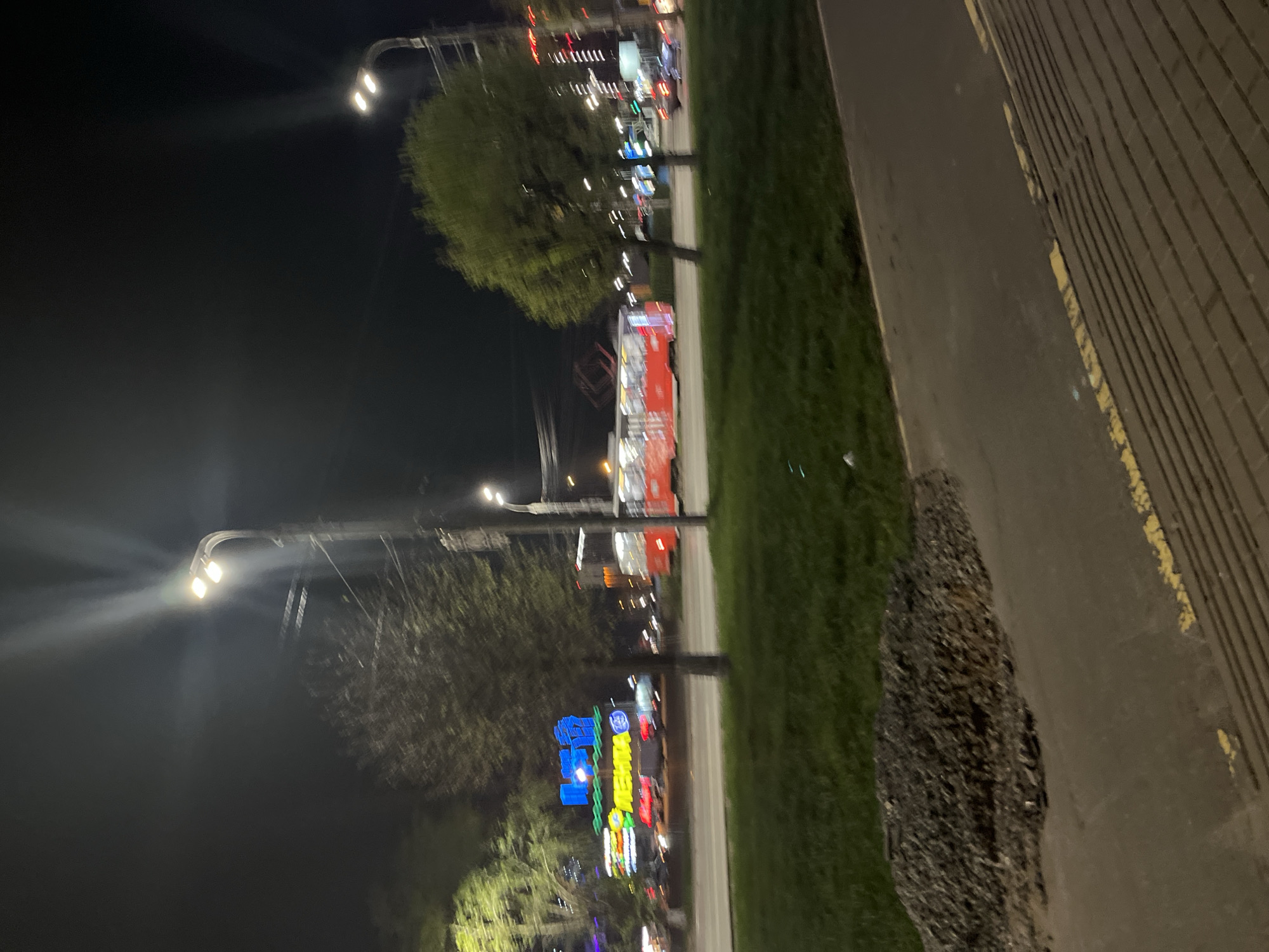 Here is a forensic description of the image:
The image depicts a nighttime urban scene along a road. It is taken at eye-level. Streetlights illuminate the area. Trees line the side of the road, and further away, buildings with brightly lit signs are visible. A wide sidewalk, edged with yellow markings, extends from the right edge of the frame, sloping slightly upwards. There's a dark, textured patch or debris on the sidewalk near the grassy area. The signs in the distance read "Petma", which is located in Izhevsk, Russia.