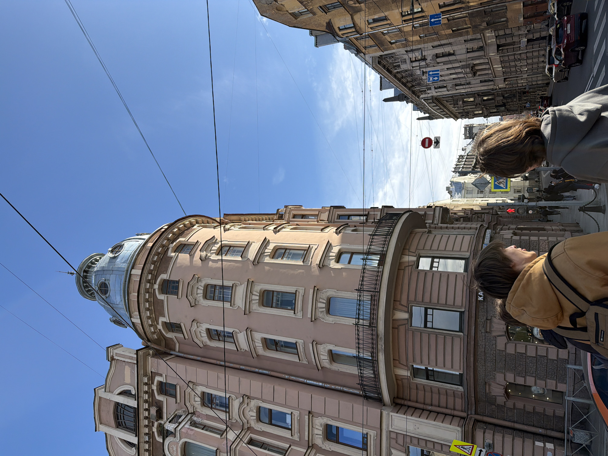 The image captures a street scene in Saint Petersburg, Russia, featuring a tall, pink-colored building with a distinctive rounded turret. The building's architecture is characterized by ornate window frames and a wrought-iron balcony. Overhead, black cables stretch across the clear blue sky. The angle of the photograph is low, emphasizing the building's height and the open sky above.
On the right side of the frame, two people stand near the viewer, their heads turned upwards as if gazing at the building. One person wears a light brown jacket and carries a shoulder bag, while the other wears a gray hooded jacket.
In the background, across the street, are other buildings with a more traditional, gray-stone facade, characteristic of Saint Petersburg's architecture. Cars are parked along the street, and street signs are visible. A red traffic light is lit. Pedestrians are walking on the opposite sidewalk. The overall impression is of a sunny day in a historic urban environment, with people admiring the architecture.