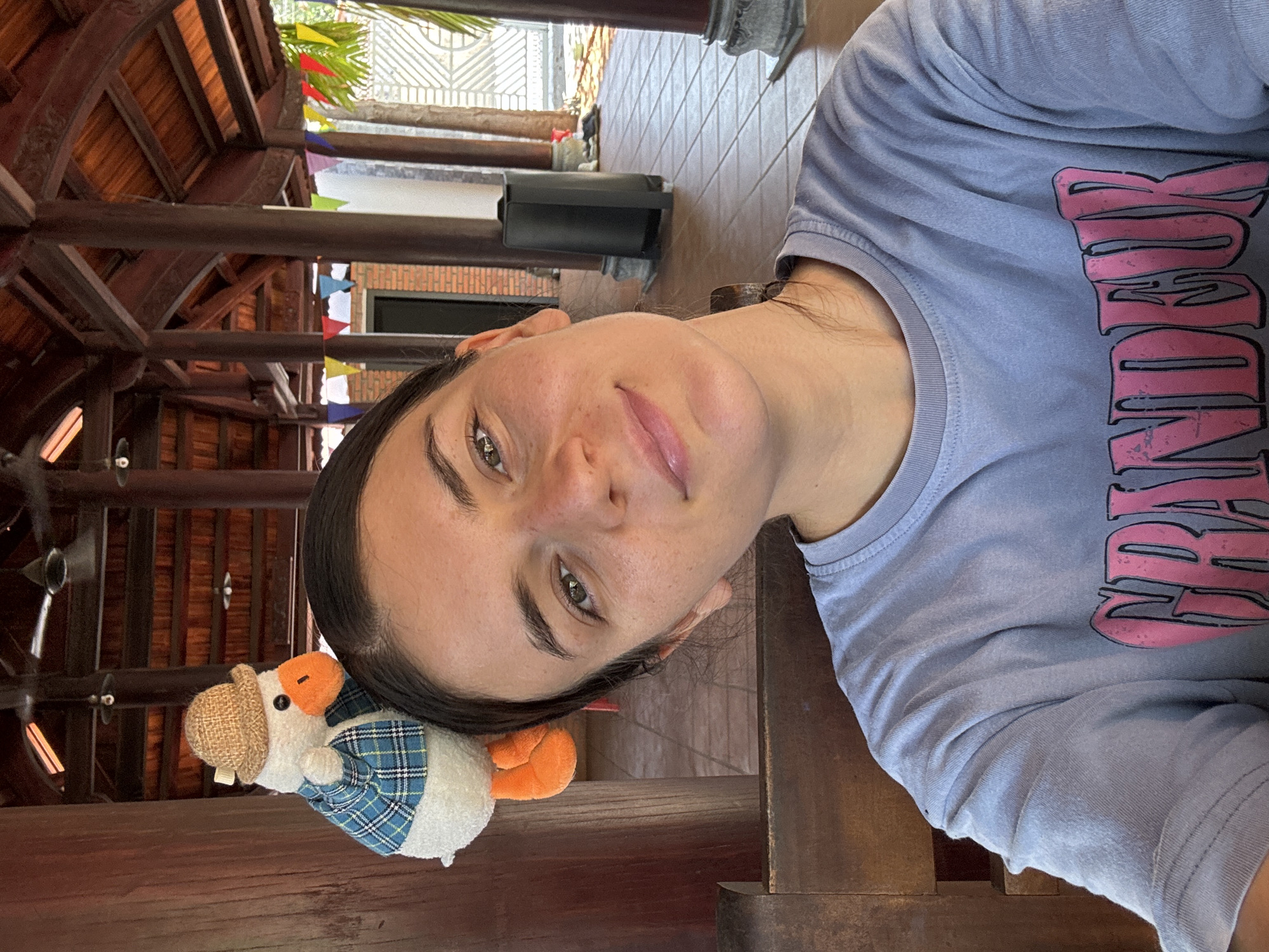 A medium shot, taken in Mũi Né, Vietnam, shows a woman and a stuffed duck. The woman, facing slightly towards the right, is wearing a light blue t-shirt with pink and black lettering. She has dark hair pulled back, light skin, and green eyes. Leaning against her head on the left is a stuffed duck wearing a brown hat and a blue and white plaid shirt. The background shows a wooden structure, possibly a gazebo or similar open-air building, with a glimpse of greenery and some colorful bunting visible outside. The floor appears to be tiled.