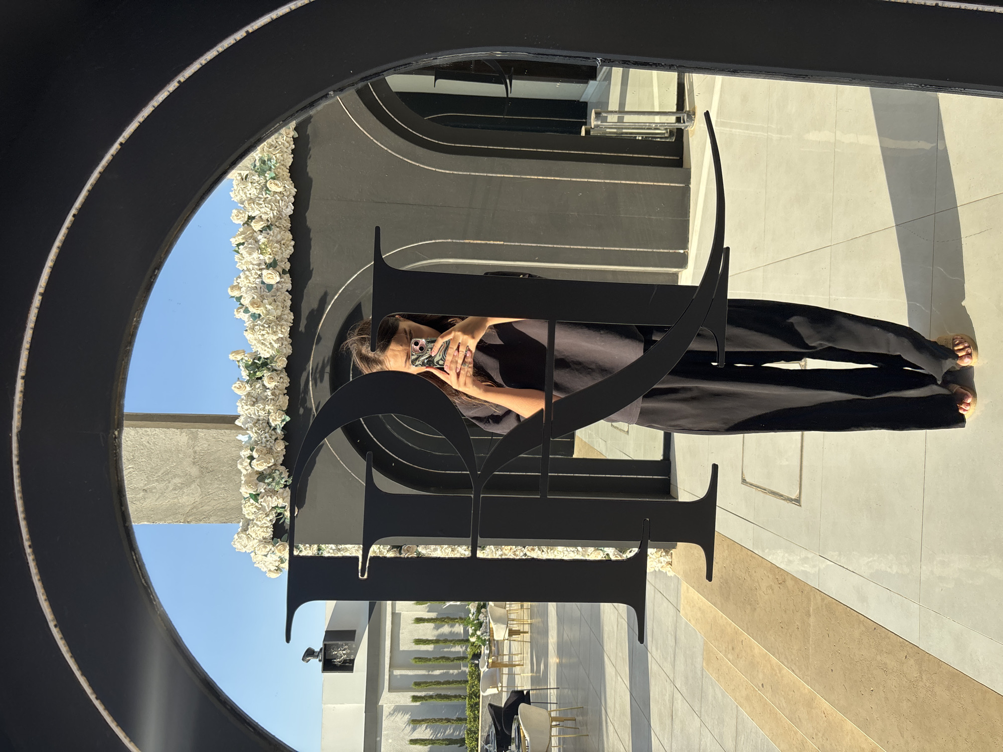 In Um Rummane, Jordan, a woman is captured in a selfie taken through a large, black, arched frame that appears to be part of a decorative installation. She is standing on a tiled patio, illuminated by bright sunlight casting distinct shadows. The woman, wearing a black t-shirt and black wide-leg pants, holds a smartphone in her hands to take the photo. In the reflection of the archway, a setup resembling an outdoor event space is visible, featuring tables and chairs, surrounded by greenery and decorative white flowers. The background includes architectural elements like columns and arched doorways.
