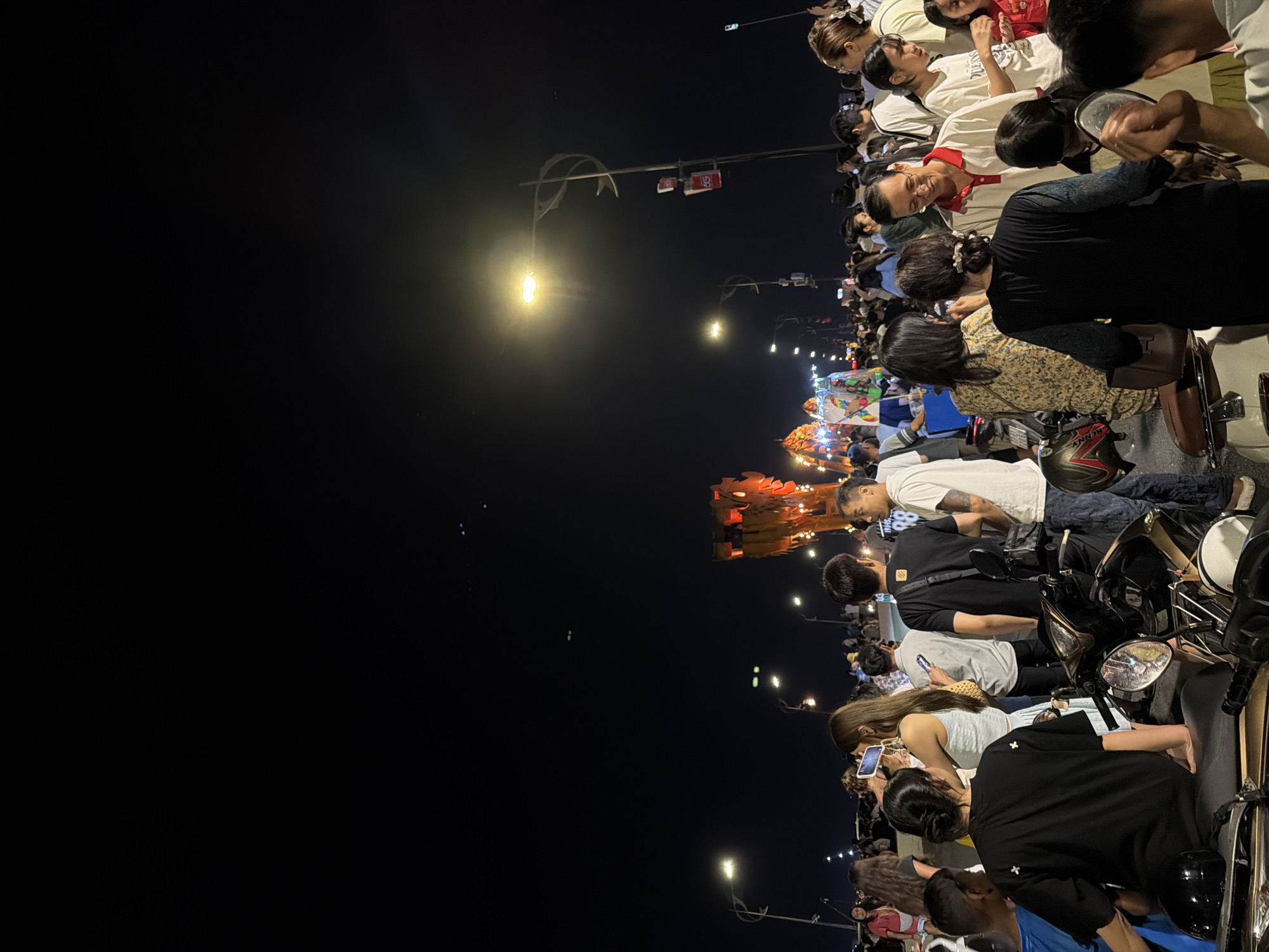 The image shows a crowd of people gathered on a street in Đà Nẵng, Vietnam, at night. Many people are on motorbikes, observing a display or event featuring a lit-up structure resembling a dragon, which is visible in the background. Some individuals are taking pictures with their phones. Streetlights illuminate the scene.