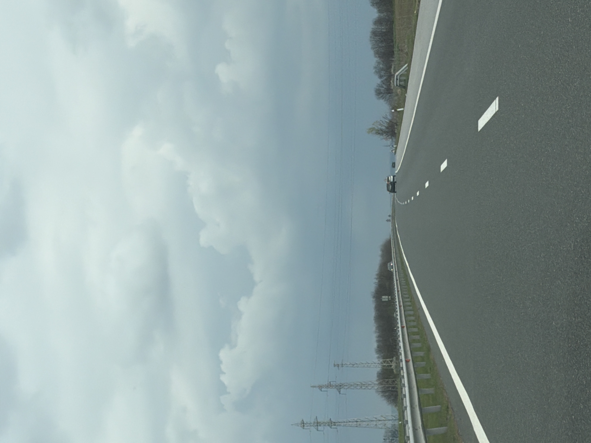 The image shows a view of a highway. The highway is made of asphalt, with white lane markings. There is a guardrail on the left side of the road. Beyond the guardrail is vegetation and power lines. In the distance, cars can be seen on the highway. The sky is cloudy. There is no evidence to suggest this is Vodenichane, Bulgaria.