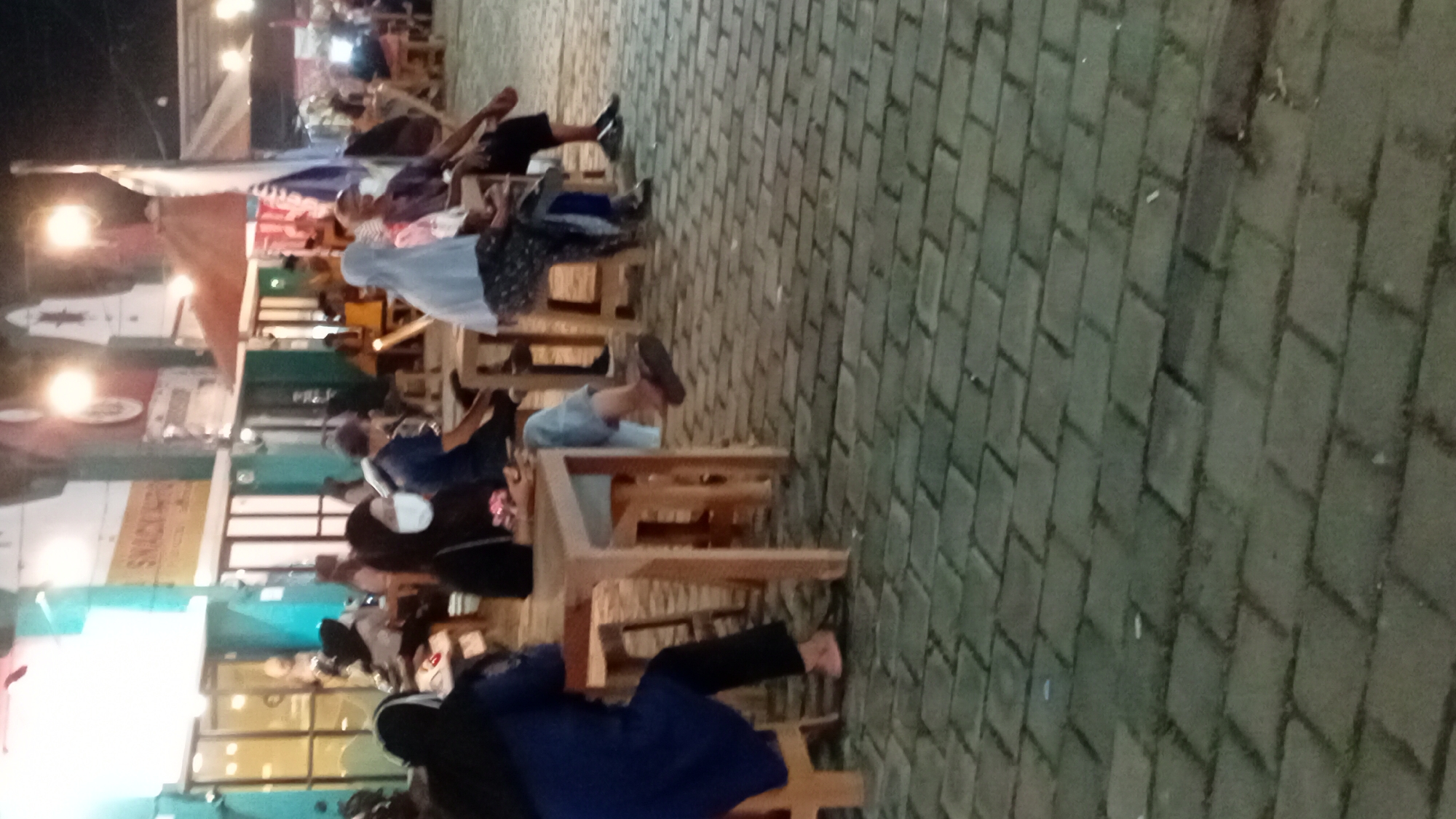This image depicts an outdoor scene at night, likely a food court or street-side dining area in Jambi City, Indonesia. Several individuals are seated at wooden tables and chairs, some facing each other, suggesting social interaction or dining. The people are dressed in casual attire, with some wearing headscarves. One person in the foreground is wearing a dark blue top and black pants, with pink flip-flops. Another person in the middle ground is wearing a white mask and a black headscarf. The ground is paved with interlocking grey bricks. In the background, there are illuminated shopfronts with signage that appears to include "SNACK INFO". Overhead lighting and umbrellas suggest a commercial area catering to evening activity. The overall atmosphere is relaxed and casual.