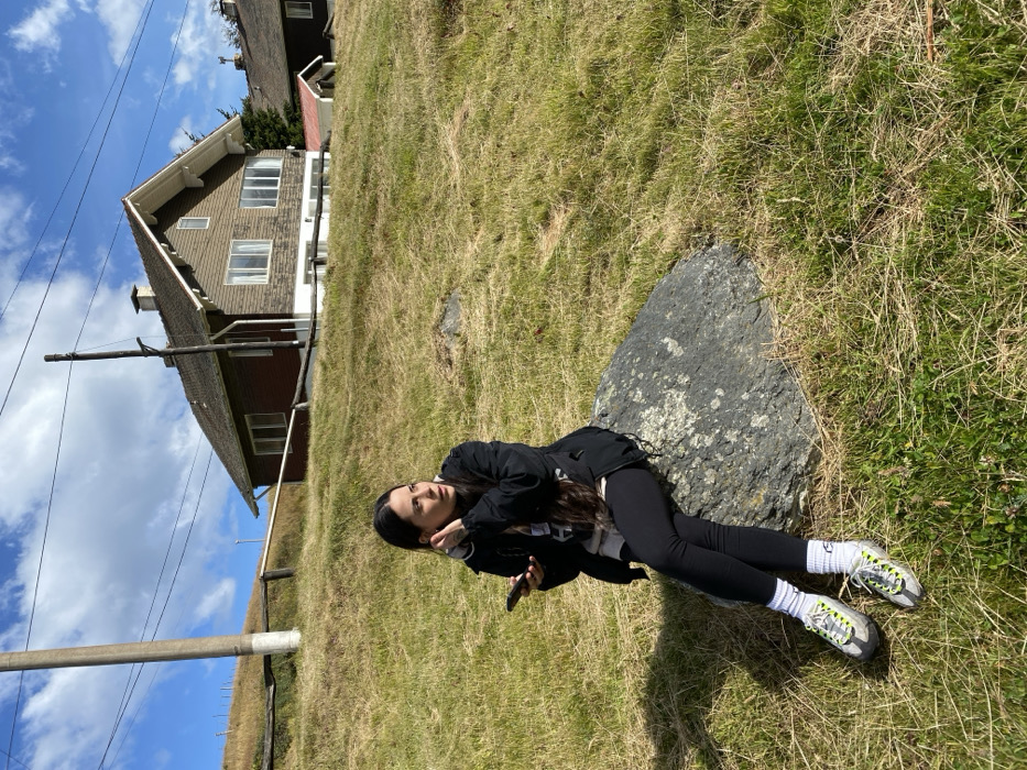 A woman in black leggings, a black jacket, white socks, and grey and green shoes reclines on a large, grey rock in a grassy field. A building is in the background, with a portion of the sky visible above it. The scene is illuminated by sunlight, casting shadows on the grass. A utility pole and wires are visible in the periphery.