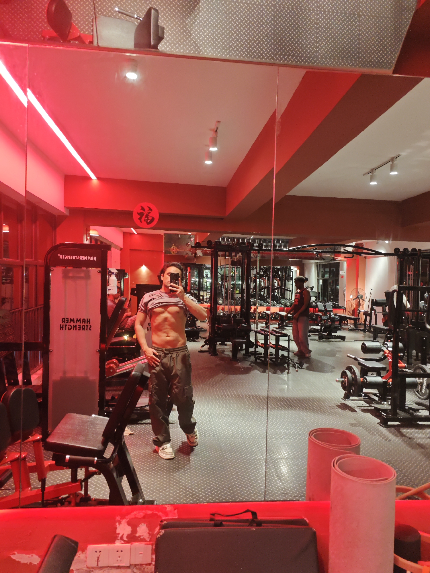 A man is taking a selfie in a gym in Chongqing High-tech Zone, China. The man has a muscular torso and is wearing casual pants and shoes. The gym has various weight machines and equipment. Another person is visible in the background. The gym is lit with red lighting. The floor is a diamond-plated pattern.
