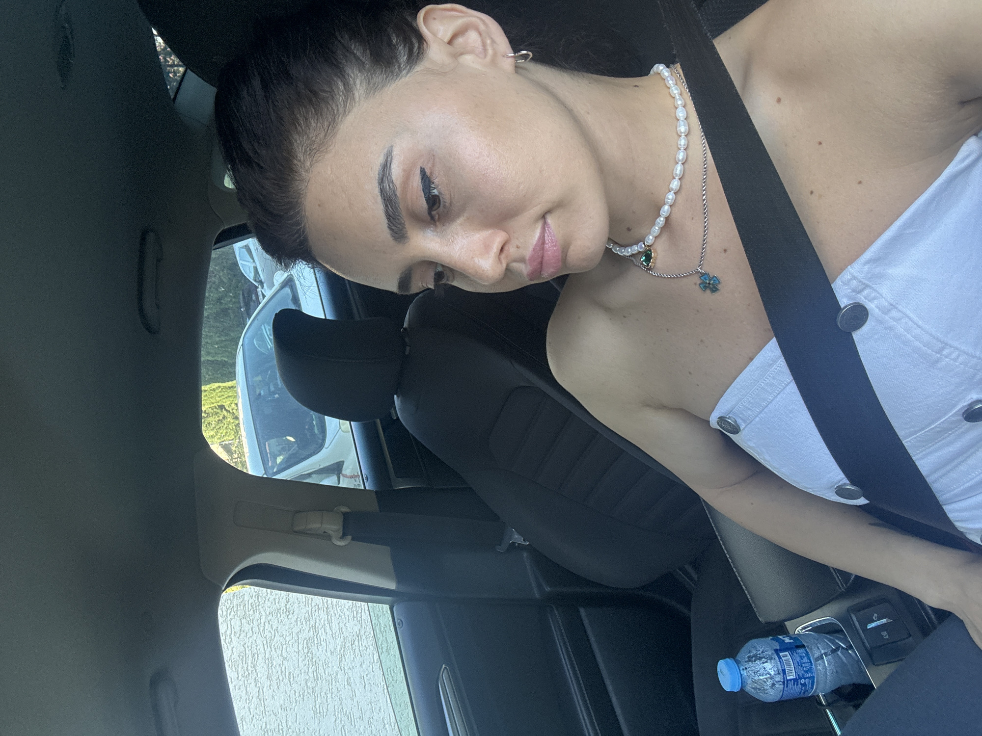 The image shows a woman sitting in the passenger seat of a car, wearing a white strapless top and a pearl necklace. The car appears to be parked on a street in Tbilisi, Georgia, as suggested by the architecture visible through the windows. The woman is looking out the window, and her face is partially obscured by shadows. A bottle of water is visible in the center console of the car. The overall lighting suggests it is daytime.