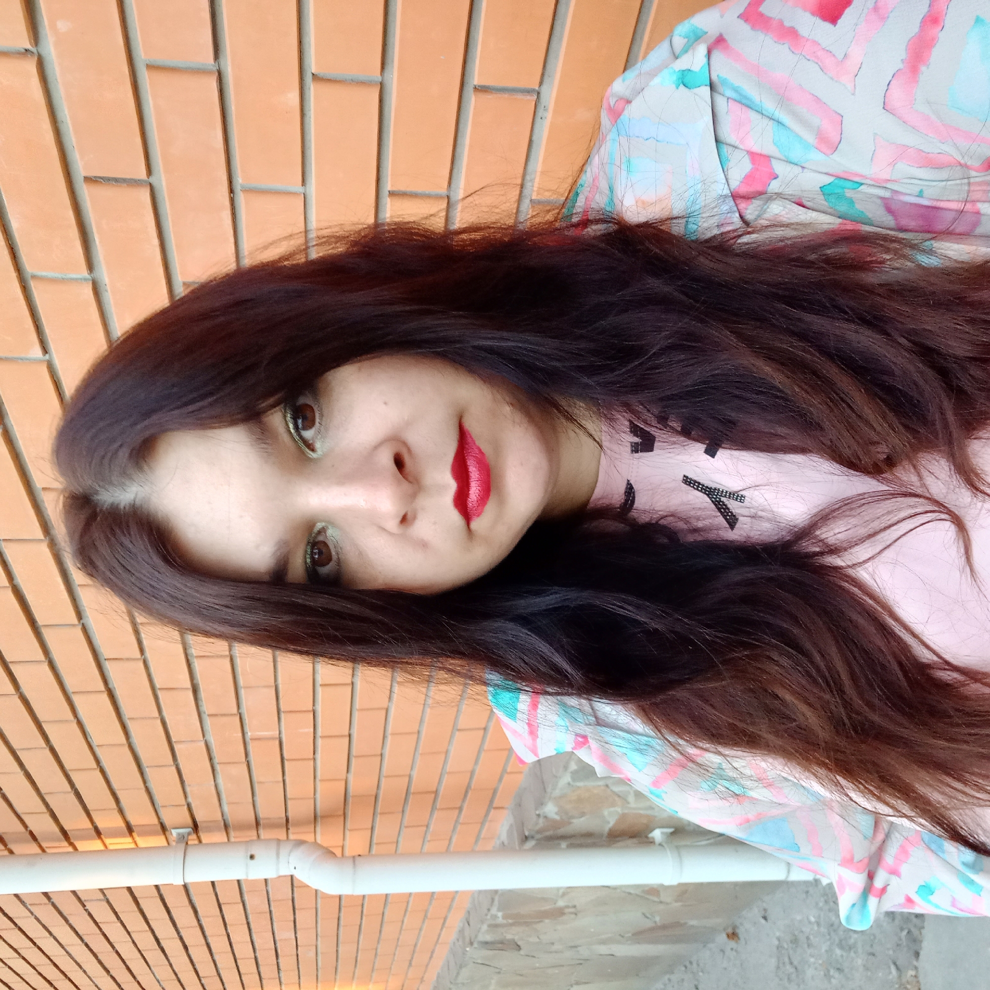 In Kropyvnytskyi, Ukraine, a young woman with long, dark hair and makeup, including red lipstick and green eyeshadow, poses for a selfie against a brick wall. She is wearing a light-colored jacket with a colorful, abstract pattern over a pink t-shirt. To her left, a white downspout runs vertically along the brick wall. The lighting suggests it is late afternoon or early evening, casting a warm glow on the scene.