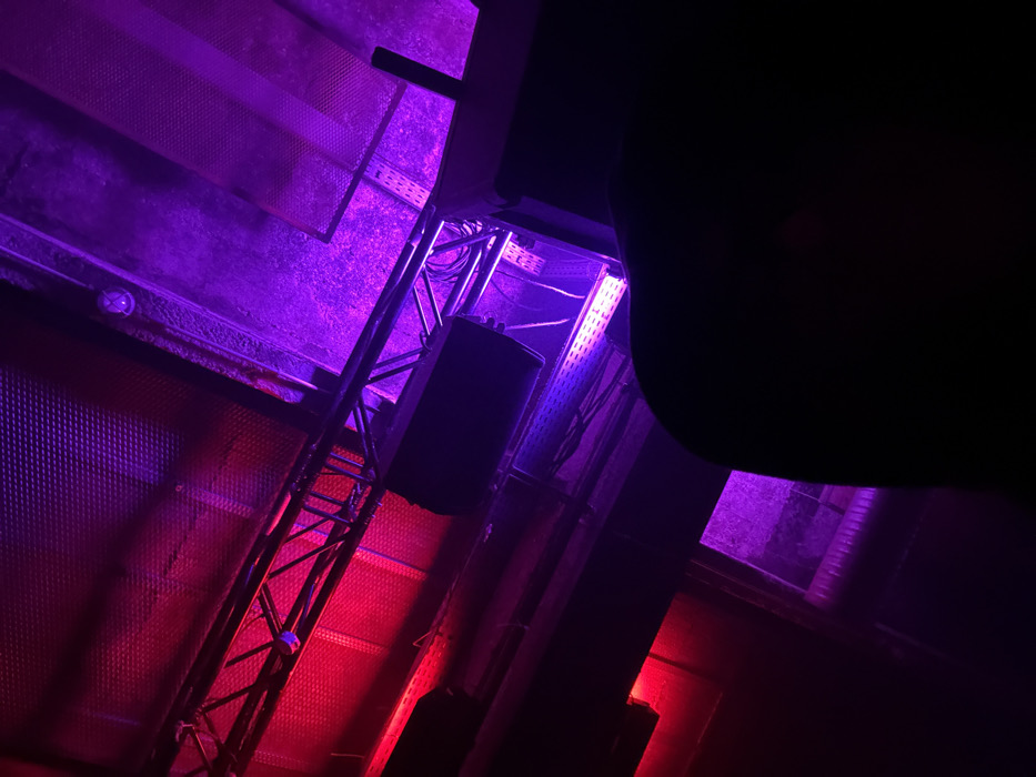 The image captures an overhead perspective inside Le Central, the main room of La Machine du Moulin Rouge, a concert hall and club located in the Montmartre district of Paris, France. The scene is bathed in dynamic red and purple stage lighting. Visible are industrial metal trusses supporting multiple large, black sound speakers and linear LED lighting fixtures. The ceiling features perforated metal panels and exposed cable trays containing wiring. The lower right portion of the frame shows the dark, indistinct silhouette of a person's head and right shoulder, suggesting a vantage point from within the audience looking upwards. No explicit actions or interactions are discernible beyond the atmospheric illumination of the venue's infrastructure.
