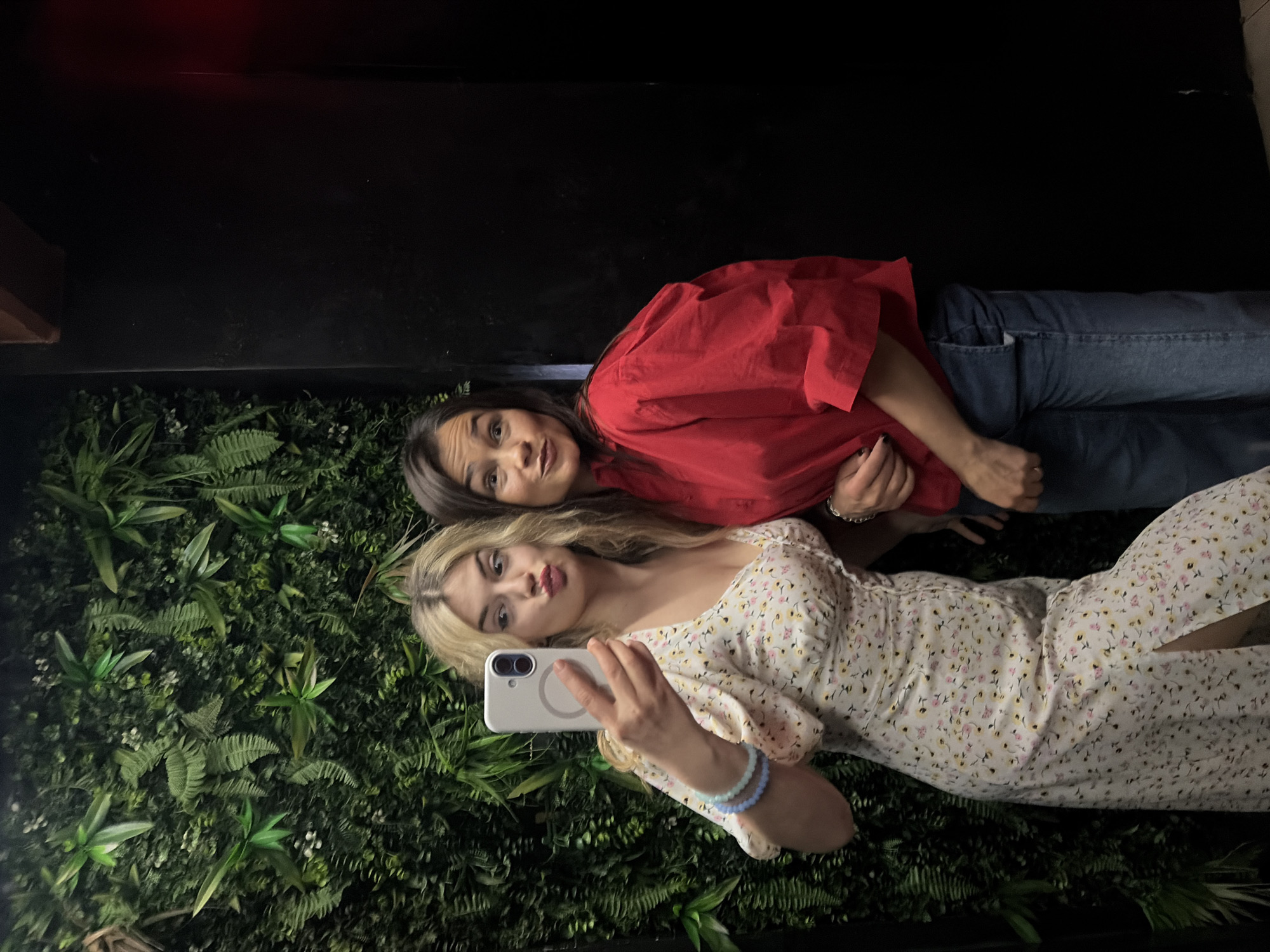 Two women are taking a selfie in Budva, Montenegro. One woman with brown hair is wearing a red shirt and jeans. The other woman has blonde hair and is wearing a floral dress. They are posing in front of a wall covered in greenery. The environment is dark, with the wall and leaves providing the only visible light source. One is holding a white smartphone.