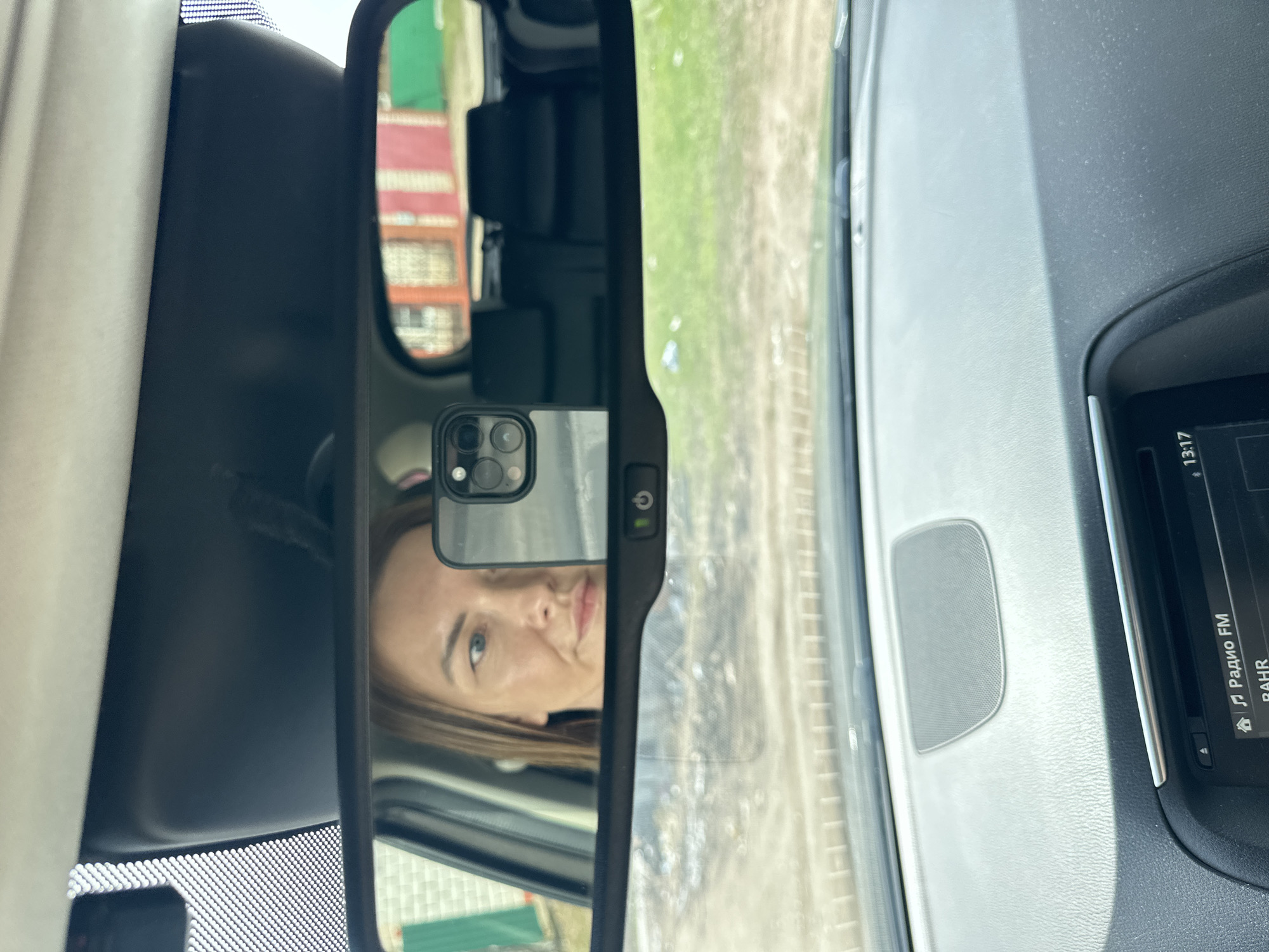 The image shows a selfie reflected in the rearview mirror of a car. The reflection captures a woman's face, holding a smartphone with a prominent camera array. The mirror also reflects a glimpse of the exterior: a building with red and green elements, and what appears to be a road or path. The interior of the car is visible, including a dashboard with air vents and an infotainment screen displaying information in Russian, including "Радио FM" (Radio FM) and the time "13:17".
While the information provided is insufficient to determine a precise location in Chaikovsky, Russia, the presence of Russian text on the infotainment screen confirms the location within Russia. Additional visual cues of the reflected structures would be required to narrow down the location within Chaikovsky.