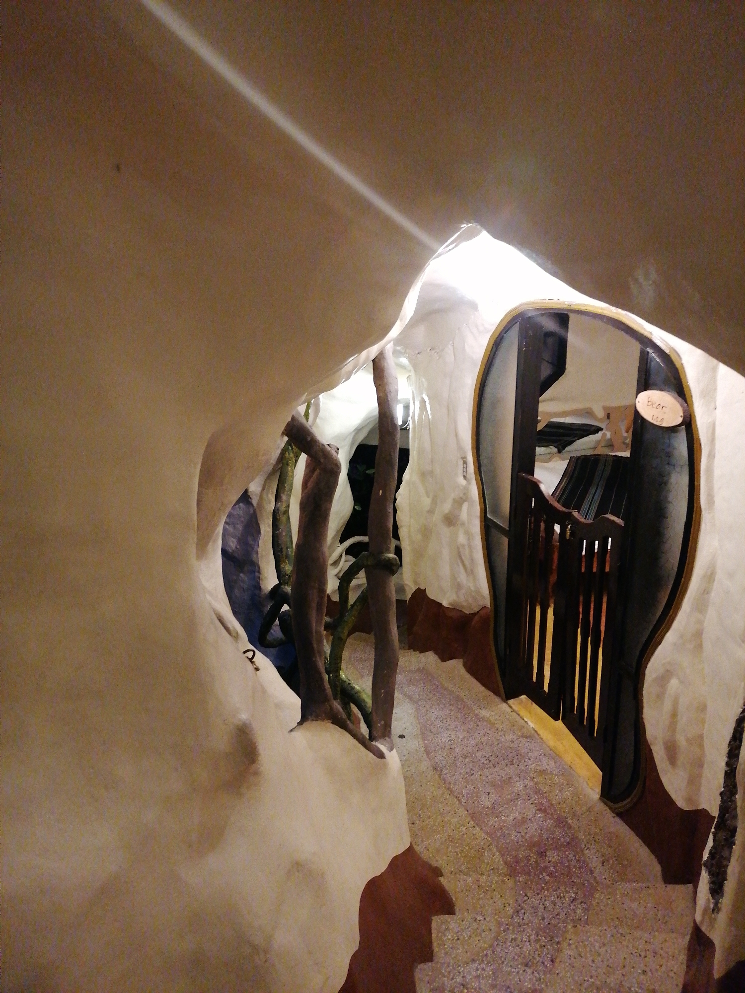 This image shows the interior of the Crazy House (Hang Nga Guesthouse) in Da Lat, Vietnam. The architecture mimics a treehouse and is known for its unconventional design. A winding tunnel-like passage is seen with beige and white textured walls. To the right, there is a door with an uneven, rounded shape and a dark wooden frame with a gold accent. The door has a dark wooden fence or barrier in front of it with vertical wooden slats. A small, oval wooden sign with the word "beer" written on it is affixed to the wall next to the door. The floor is a speckled mix of colors and appears to be made of a terrazzo-like material. Natural-looking wooden supports are visible throughout the space.