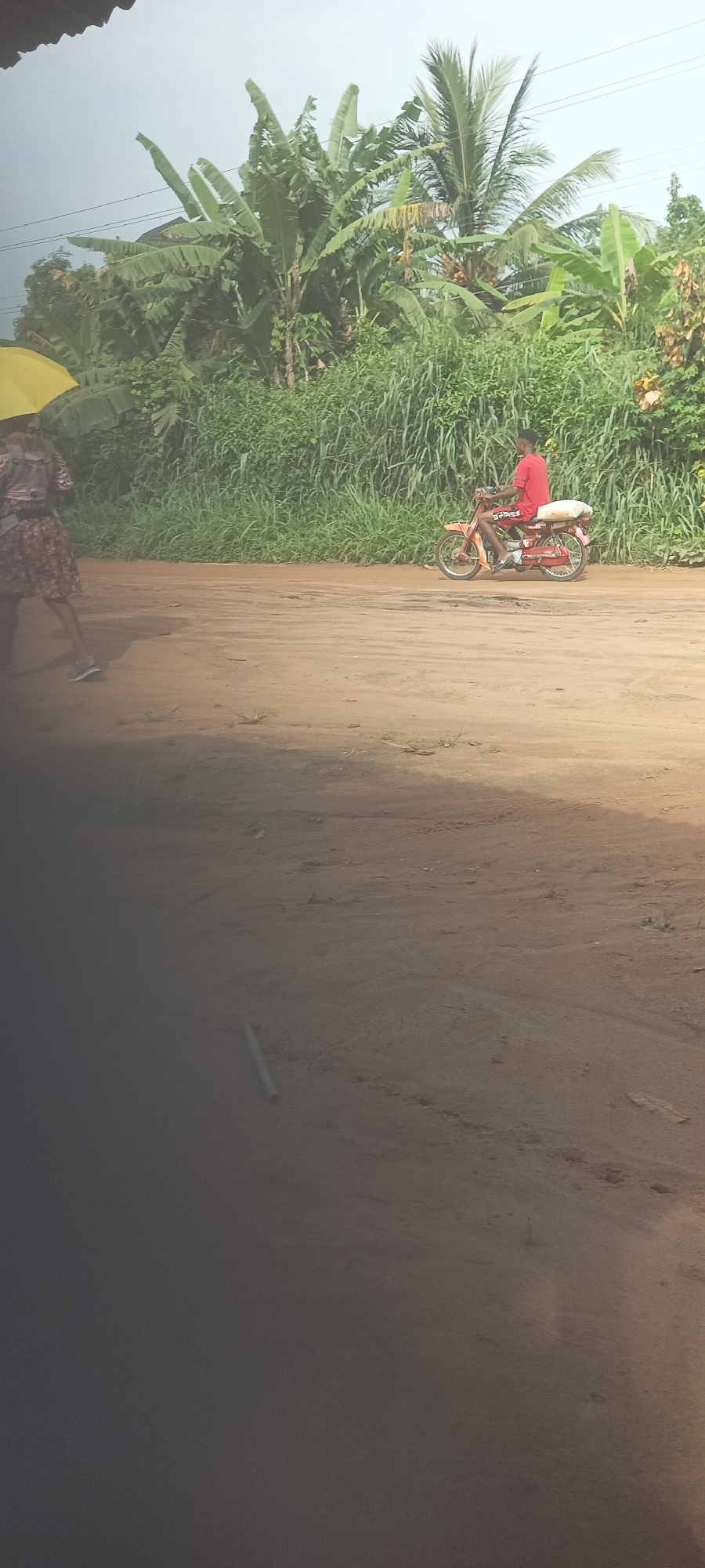 The image depicts a dirt road in Amauzu, Nigeria, flanked by dense vegetation including banana trees and tall grass. A person is riding a red motorcycle along the road, carrying a white object on the back. To the left, a person holding a yellow umbrella is walking. The foreground is obscured by shadows, suggesting the photo was taken from an indoor location or shaded area.