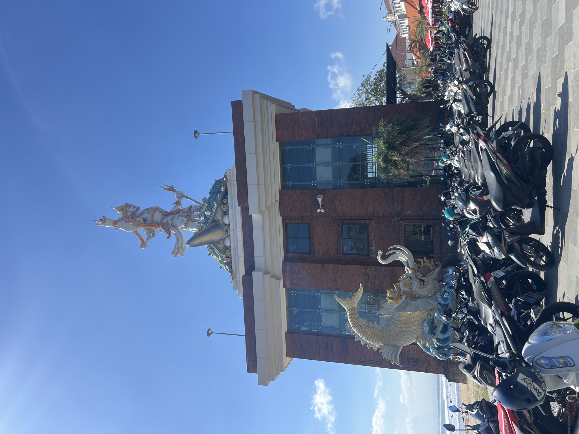 This is a photograph taken in Kuta, Indonesia, featuring a large monument atop a brick building. The monument depicts a multi-armed deity holding weapons, with a mythical sea creature, resembling a dragon or a large fish, at its base. The building has large glass windows and a white decorative facade at the top. In front of the building, a large number of motorcycles are parked on a paved area. Beyond the building and motorcycles, a beach is visible with people in the ocean, suggesting a coastal location. The sky is clear blue with a few wispy clouds. The overall scene suggests a vibrant, public space in a tropical urban environment.