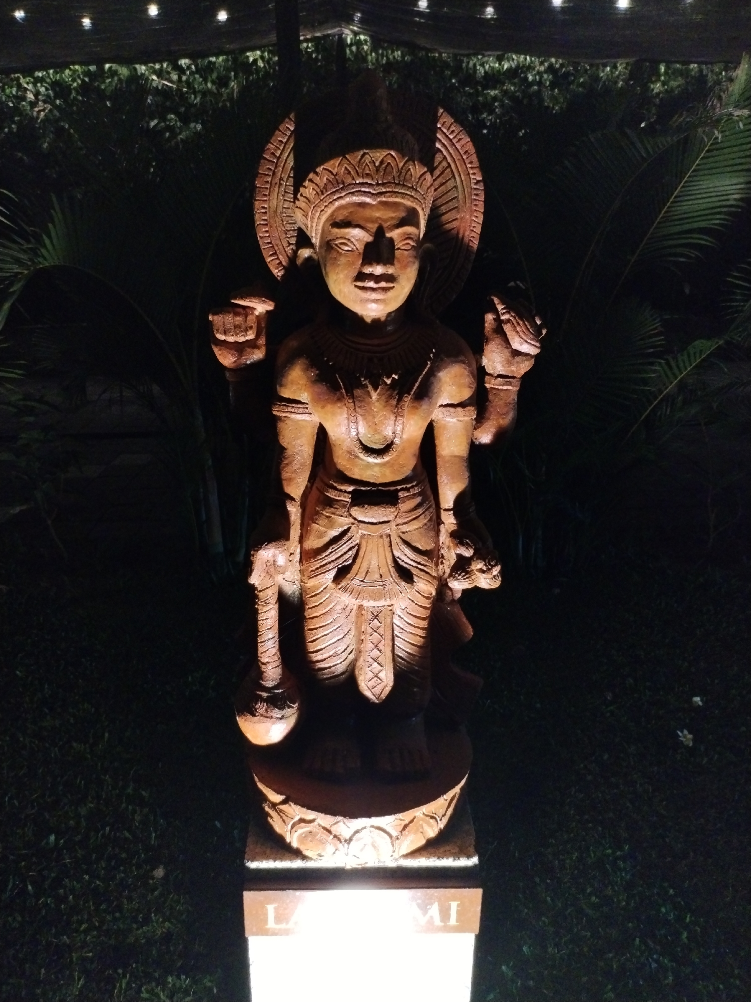 The image shows a statue of an Asian deity standing in what appears to be a garden at night. The statue is made of a reddish-brown material, likely stone or wood, and features four arms. The deity is adorned with a headdress and jewelry. The statue is illuminated from below, casting dramatic shadows. The background is filled with dark foliage, with a few palm-like leaves visible. Overhead, there are faint points of light, perhaps from string lights. The statue is set on a square base with lettering that appears to read "LAXMI." The statue is located at Bac Nha Trang Ward, Vietnam.