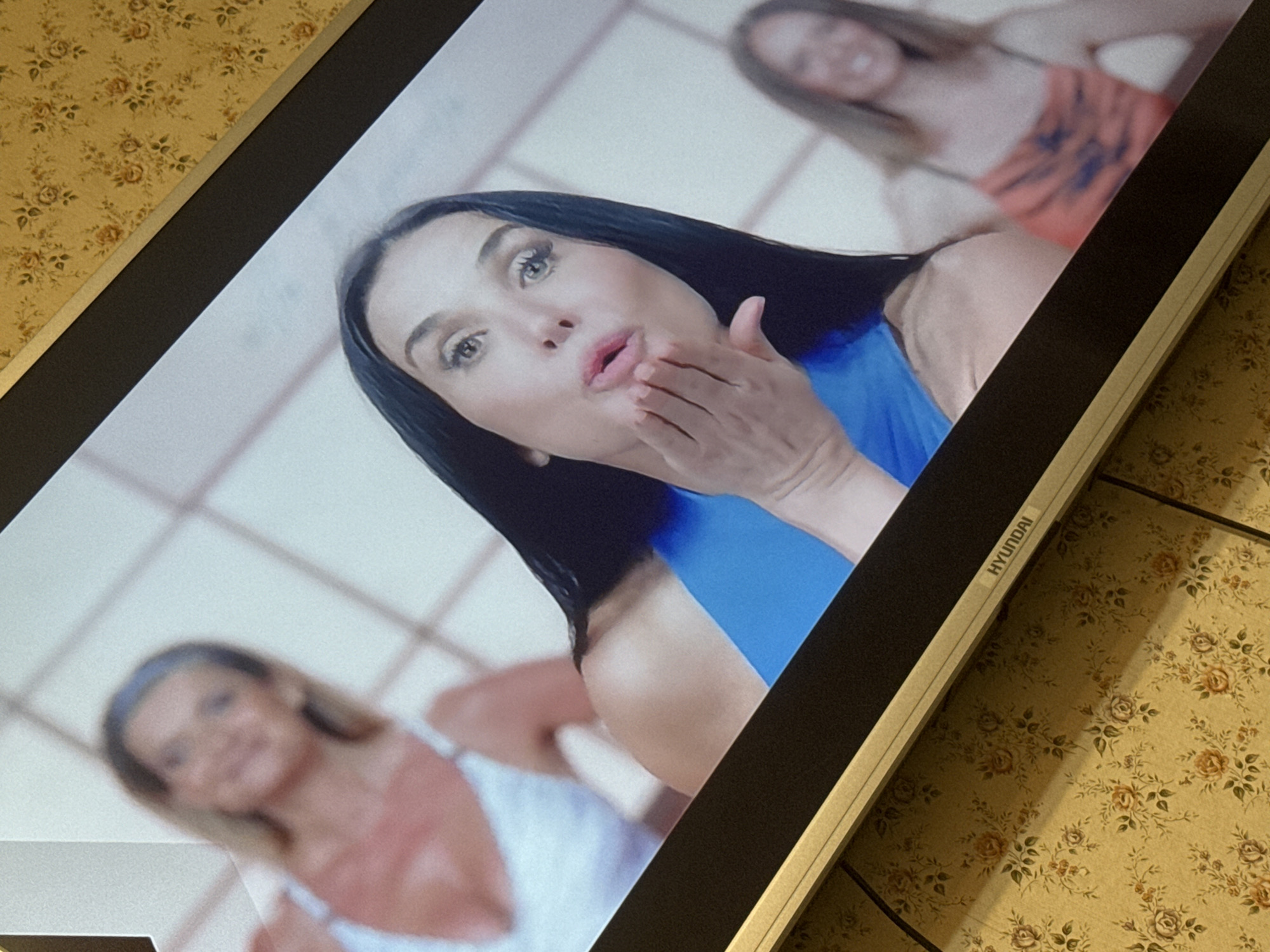 A close-up shot depicts a Hyundai television screen displaying a scene with three women. The primary focus is on a woman with dark hair and fair skin who is puckering her lips as if blowing a kiss. She is wearing a blue top. Two other women are visible in the background, blurred and less defined. One is wearing a white top and headband, while the other is wearing an orange top. The scene appears to be set in an indoor location with white walls. The TV is placed against a surface with a patterned wallpaper of small flowers in shades of brown, beige, and green. The location is not discernible from the image, and cannot be identified as being in Mytishchi, Russia.