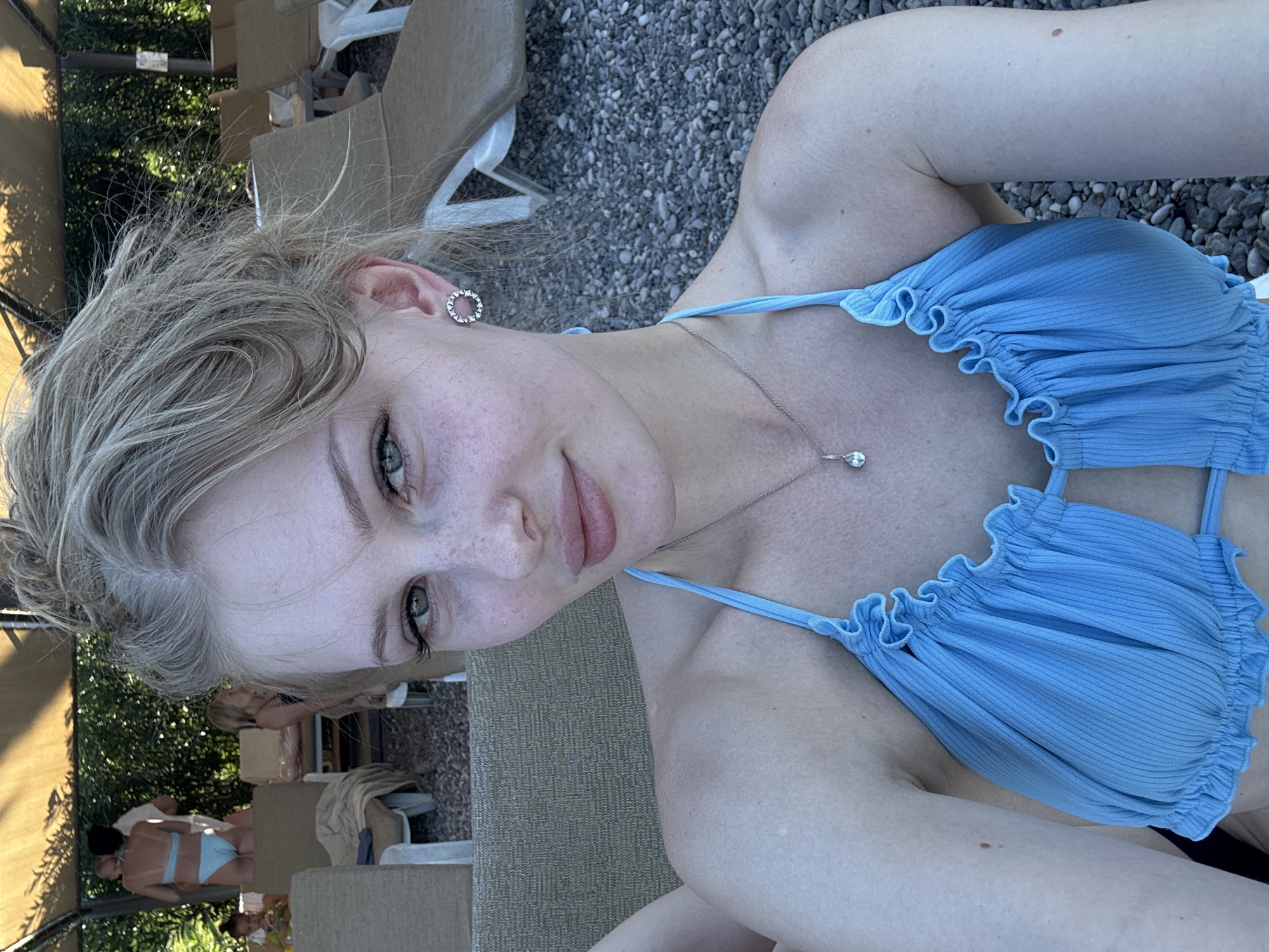 A young Caucasian woman with blonde hair is taking a selfie outdoors, possibly at a beach resort. She is wearing a light blue bikini top with ruffled edges and a silver pendant necklace. She has freckles and green eyes, and her hair is pulled up in a messy bun. In the background, several lounge chairs are visible, some occupied by other people, and dense green foliage can be seen behind them, suggesting a resort environment. The ground appears to be covered in pebbles. The overall impression is of a casual vacation setting, likely during daylight hours, consistent with a location like Kemer, Turkey, which is known for its beach resorts and pebble beaches.