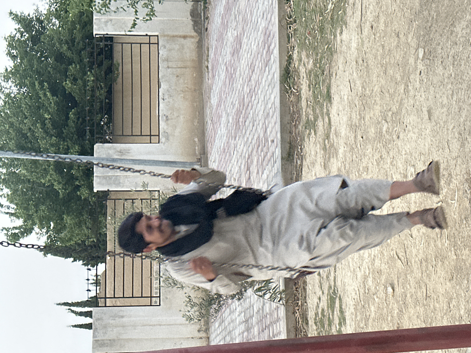 A man is depicted mid-swing on a metal swing set, captured in a moment of motion. He has dark hair, a beard, and a mustache, and is smiling or smirking, suggesting enjoyment. He is dressed in a light-colored, possibly traditional, long garment, over which he wears a dark jacket or vest. His feet, clad in light-colored shoes, are off the ground, indicating the height of his swing.
Behind the man, there is a patterned brick-paved area, likely part of a walkway or ground cover. Beyond this, a light-colored concrete wall with a rectangular opening protected by a black metal grille or fence is visible. Lush green trees with dense foliage are seen peeking from behind and above this wall. To the right of the paved area, the ground transitions to dry, sandy soil with sparse, light-colored vegetation. A red horizontal bar, possibly part of another structure, is partially visible in the bottom foreground.
The scene is outdoors, under bright, diffuse lighting, suggesting daytime, possibly under an overcast sky or during a period without direct harsh sunlight. There are no strong shadows, contributing to the soft illumination. The overall setting appears to be a park, playground, or a residential courtyard. No visible text is present in the image.