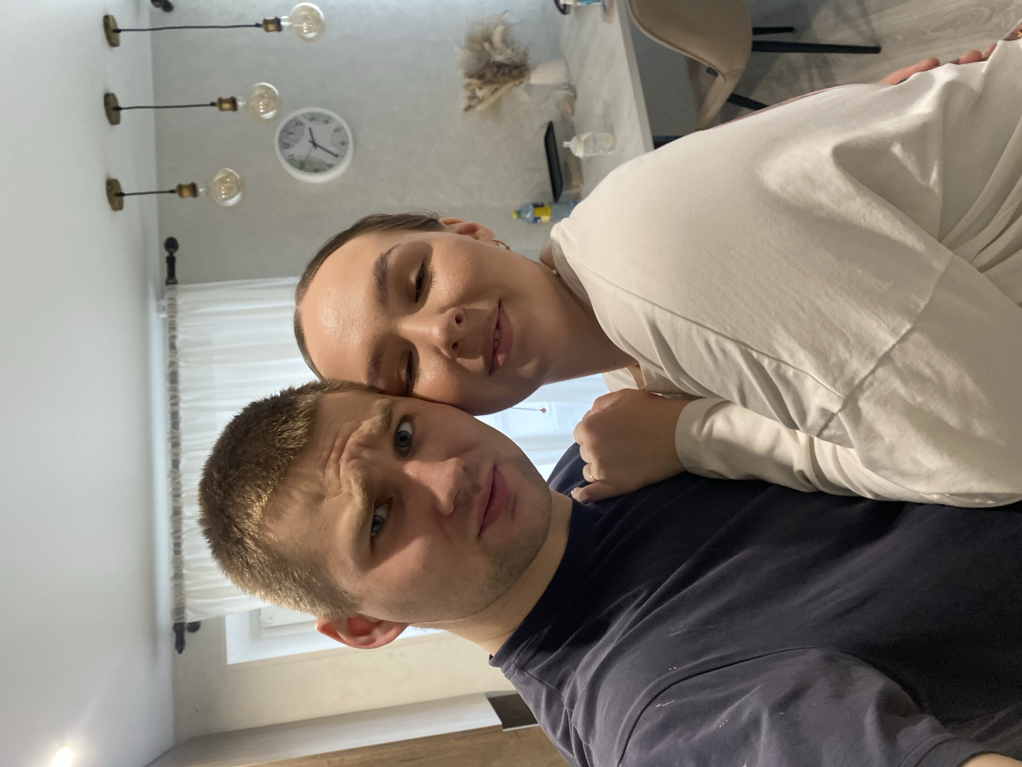 A selfie captures a moment between a man and a woman in an indoor setting, likely a home in Chaikovsky, Russia. The image is rotated counter-clockwise.
The woman, positioned closer to the camera and leaning into the man, has light skin and appears to have dark hair pulled back. She wears a light-colored (possibly white or off-white) long-sleeved top and exhibits a soft, contented smile with her eyes partially closed, resting her head on the man's shoulder or back.
The man, with light skin and short light brown hair, wears a dark blue or navy t-shirt which has some light marks or stains on it. He looks directly at the camera with a slightly quizzical or amused expression, his eyebrows raised. His left arm is visible, with a wedding band on his finger, suggesting the two are a married couple.
The background reveals a domestic environment with light-colored walls. Three industrial-style light fixtures with exposed filament bulbs are mounted on the wall or ceiling. A white circular wall clock with black hands and numbers indicates approximately 1:55. Behind the woman, a light-colored table holds a dried floral arrangement, a mobile phone, and a water bottle. A beige chair is partially visible near the table. Behind the man, a window with sheer white curtains is visible, suggesting daylight is entering the room.
The overall mood is intimate and relaxed, capturing an affectionate interaction between the two individuals.