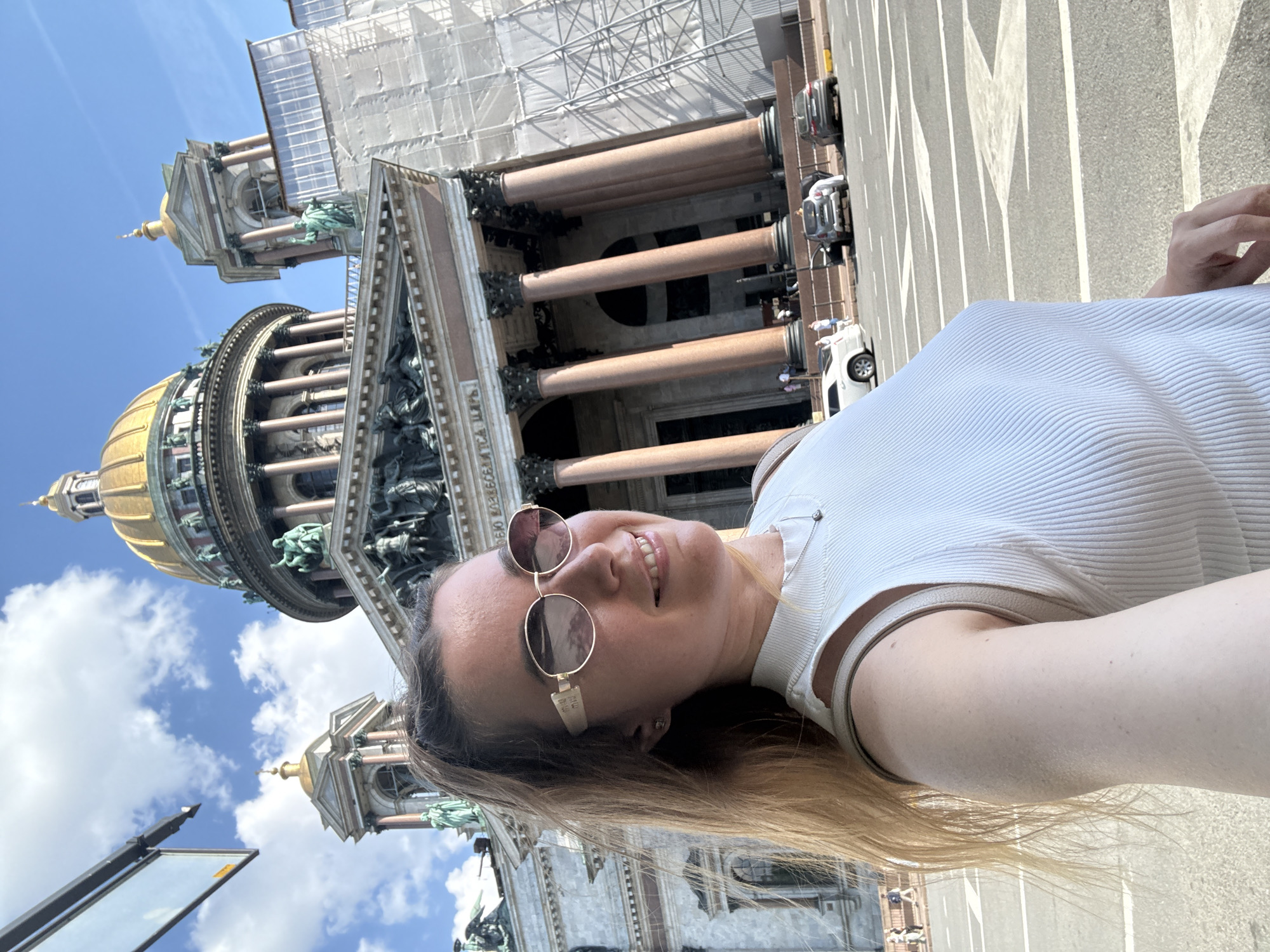 The image is a selfie taken by a smiling woman, rotated 90 degrees counter-clockwise, with a grand architectural landmark in the background. The woman has long, light brown hair and is wearing a white ribbed top and round, reflective sunglasses. She is looking towards the camera with a wide smile, her face illuminated by bright sunlight.
Behind her, a massive, ornate building with classical architectural elements dominates the scene. It features large, reddish-brown columns, intricate sculptural details, and a prominent golden dome. Part of the building's facade, particularly towards the upper right, is covered in extensive scaffolding, indicating ongoing construction or renovation. The structure strongly resembles Saint Isaac's Cathedral in Saint Petersburg, Russia. Below the scaffolding, some partially visible Cyrillic text can be discerned on the building's facade, with parts of the word "ВСЕХ" being somewhat legible.
The sky is clear and blue with scattered white clouds, suggesting a sunny day. In the background, a street runs alongside the building, with several cars parked or driving on it, and white road markings are visible on the asphalt. The perspective indicates the photo was taken from a low angle, looking up at the towering building. The overall impression is that of a person enjoying a visit to a significant historical or religious site during pleasant weather.