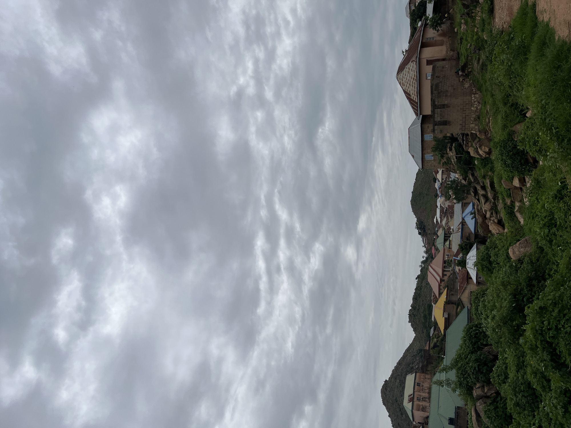 The image depicts a residential area in Jos, Nigeria, characterized by a collection of houses situated on a hilly terrain under a cloudy sky. The architecture of the houses varies, with some featuring corrugated metal roofs in colors such as green, yellow, and brown, while others have concrete or tiled roofs. The buildings are constructed from materials like brick and stone. Dense green vegetation, including bushes and trees, covers the landscape, interspersed with large rocks. The overall environment suggests a rural or semi-urban setting within Jos.