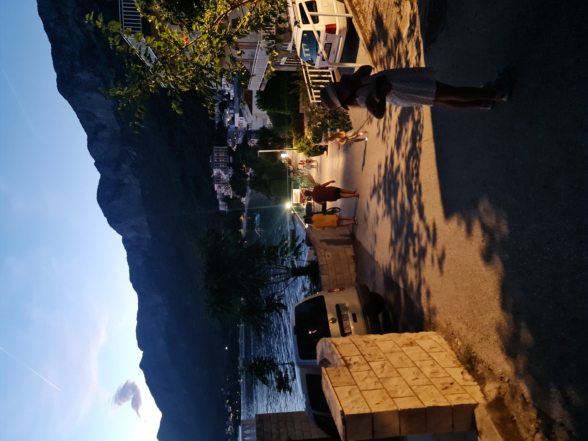 The image depicts a twilight scene on a paved road in Općina Gradac, Croatia. In the foreground, a young girl wearing a white dress and a hat stands on the right side of the road, facing away from the camera and appearing to hold something to her ear. Further up the road, several people are walking: a person in a yellow shirt and shorts, another in a red shirt and shorts, and two individuals in the distance. A white van is parked on the right side of the road, and a silver van is partially visible on the left. The road is lined with a stone wall on the left and a building with a balcony and a tree on the right. In the background, a body of water, likely the Adriatic Sea, is visible, with a few buildings and lights along the shore and a large mountain range rising behind them. The sky is a deep blue with a hint of pink and a faint contrail from an aircraft. The lighting suggests the sun has recently set.