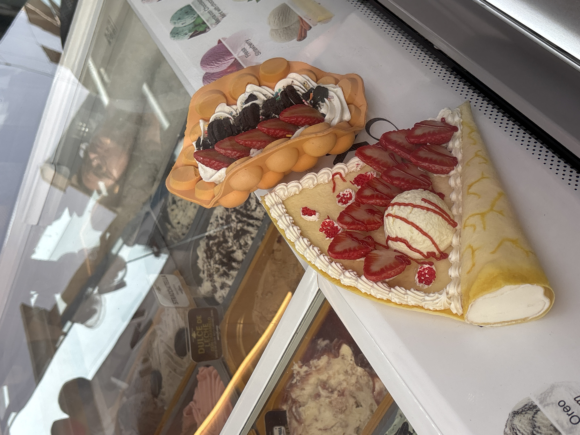 This image shows a close-up of a display case filled with various ice cream flavors and two ice cream desserts. The desserts are a bubble waffle cone topped with strawberries, whipped cream, and chocolate cookies, and a crepe folded into a cone shape, filled with ice cream, strawberries, and whipped cream. The ice cream flavors visible in the display case have labels with Spanish text such as "DULCE DE LECHE CON CARAMELO" (Dulce de leche with caramel) and "STRACCIATELLA." There is also a label for "Oreo" ice cream. The overall setting appears to be an ice cream shop or parlor, likely in Orihuela, Spain, given the context. The lighting suggests it could be daytime. The image is taken from a slightly high angle, looking down into the display. Reflections on the glass of the display case are visible, showing what appears to be a person with glasses. The desserts are artfully arranged and appear to be for display rather than immediate consumption.