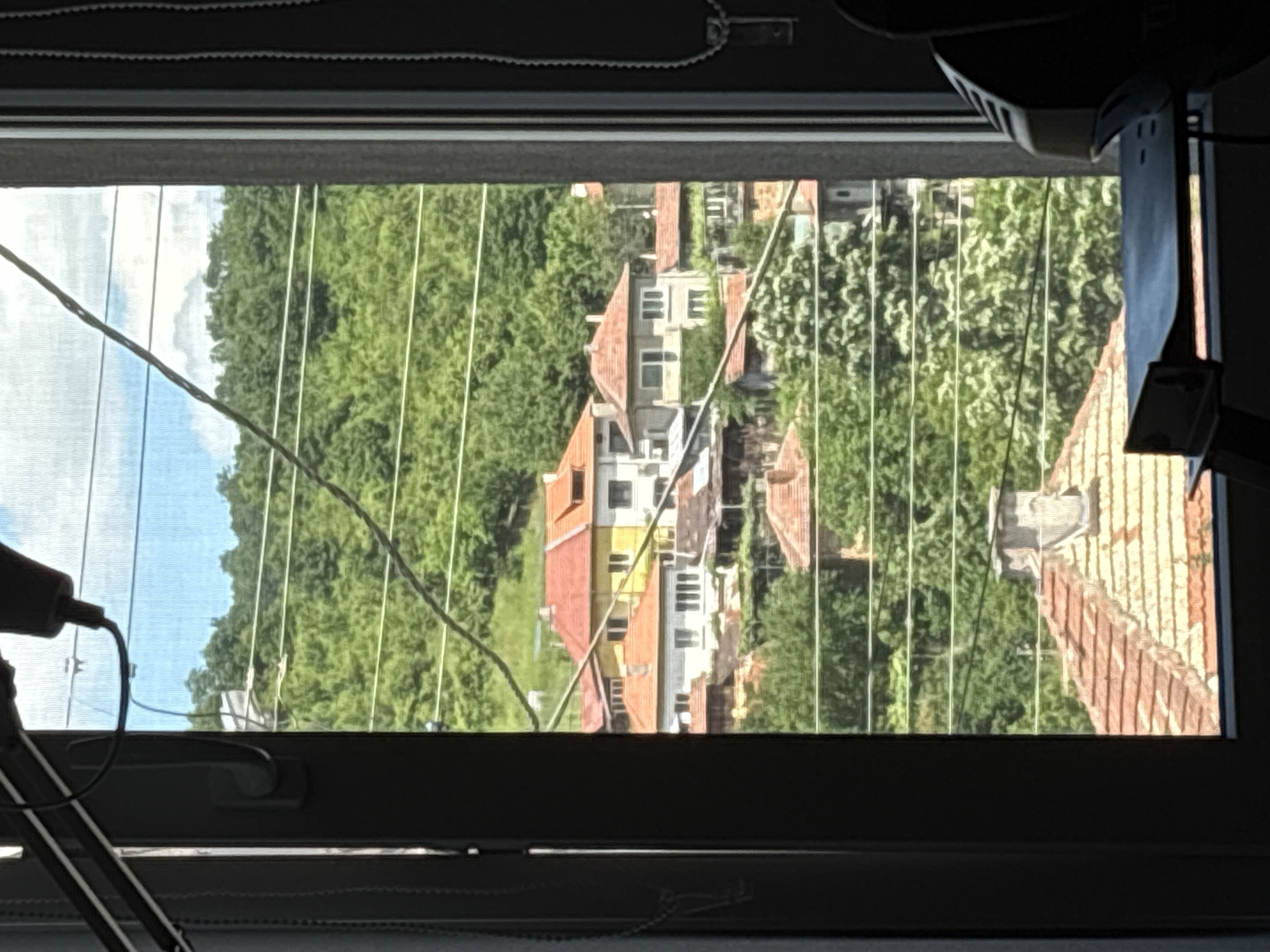 The image, taken from inside a room looking out a window, shows a view of houses and lush green hills under a partly cloudy sky. The window frame, a dark grey, partially obscures the scene. Several thin wires stretch horizontally across the view, creating a layered effect. In the mid-ground, several houses with red-tiled roofs are visible. One prominent house has a bright yellow facade with white trim and red roof. Other houses are painted in white and cream colors, also with red roofs. The background is dominated by a densely forested hillside. The foreground shows parts of the interior of the room, including a dark object, possibly a piece of furniture, and what appears to be a desk with a black object on it. The overall impression is of a residential area nestled within a natural, hilly landscape, typical of the outskirts of Gabrovo, Bulgaria.