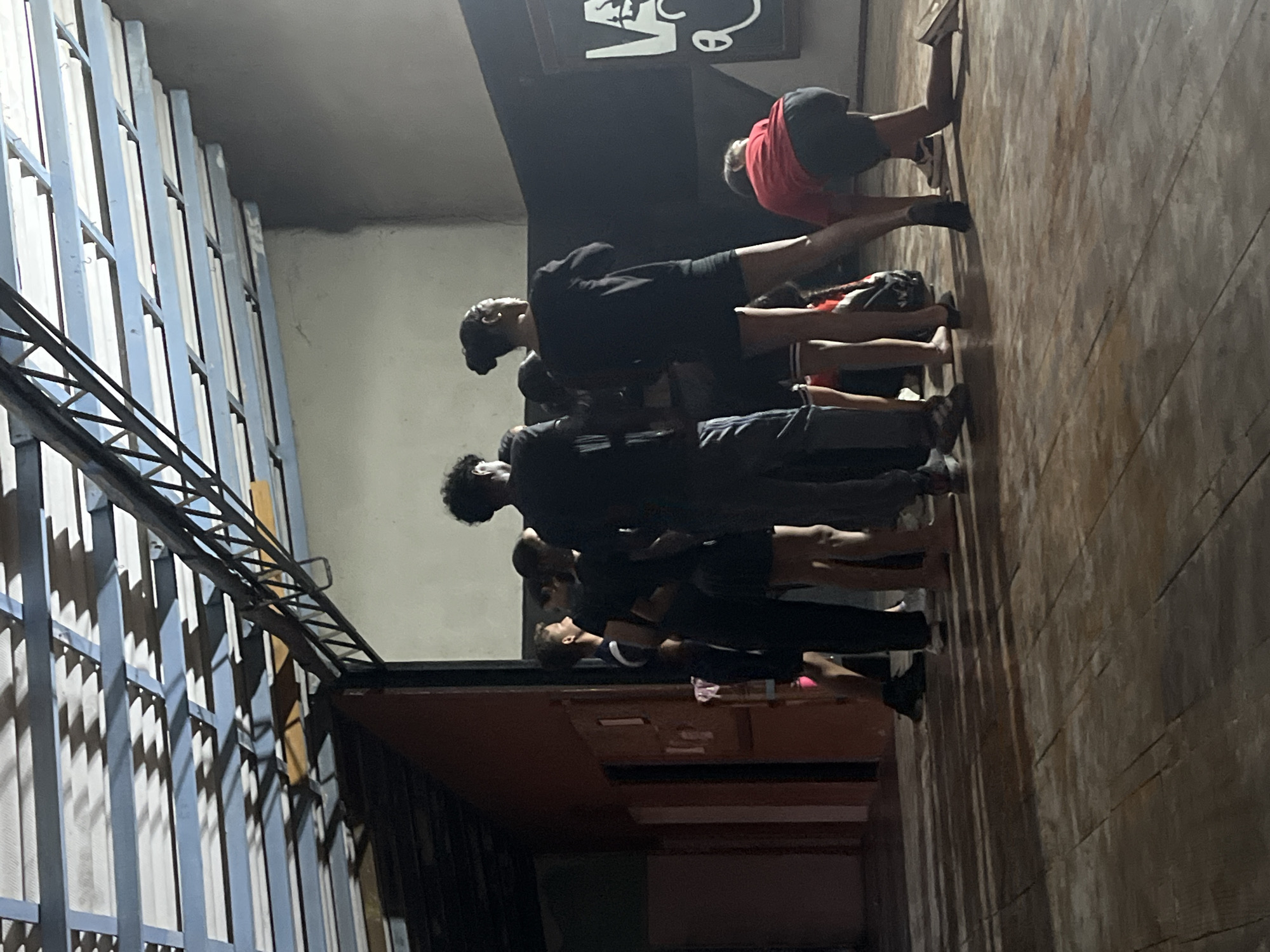 A group of approximately seven individuals are lined up on a dark-stained wooden floor in a dimly lit space, with some individuals facing and touching a lighter-colored wall behind them. At the top of the frame, an opening is visible, indicating an outdoor area. A metal grate is positioned along the side, and the scene is in Juan Griego, Venezuela.