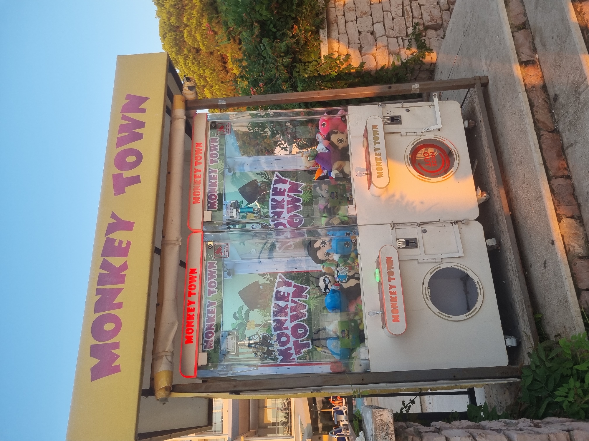 The image shows two claw machines labeled "MONKEY TOWN" in the city of Bar, Montenegro. The machines are white with a yellow awning displaying the words "MONKEY TOWN" in purple letters. Each machine has a clear glass front displaying stuffed animals. The machines are located outdoors, with a stone wall and green foliage in the background. The sun is setting, casting a warm glow on the scene. There are no people visible in the image.