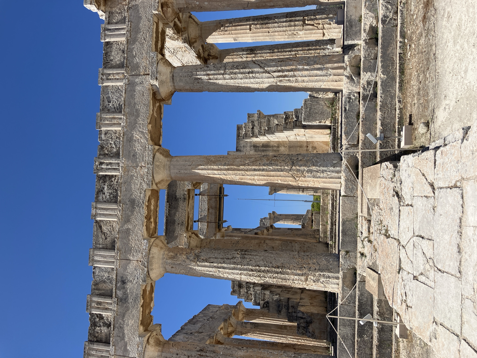 The image captures a view of ancient Greek ruins under a clear blue sky. The foreground consists of large, worn stone steps and paved areas, indicative of a historical site. Dominating the frame are massive stone columns, some intact and supporting a partial entablature, while others are broken or missing. The architecture suggests a temple or similar monumental structure. In the background, more columns and crumbling walls are visible, hinting at a larger complex. There are no people visible in the image. The setting is likely an outdoor archaeological site, potentially on a sunny day given the bright lighting and clear sky. This ancient temple is located in Mesagros, Greece.