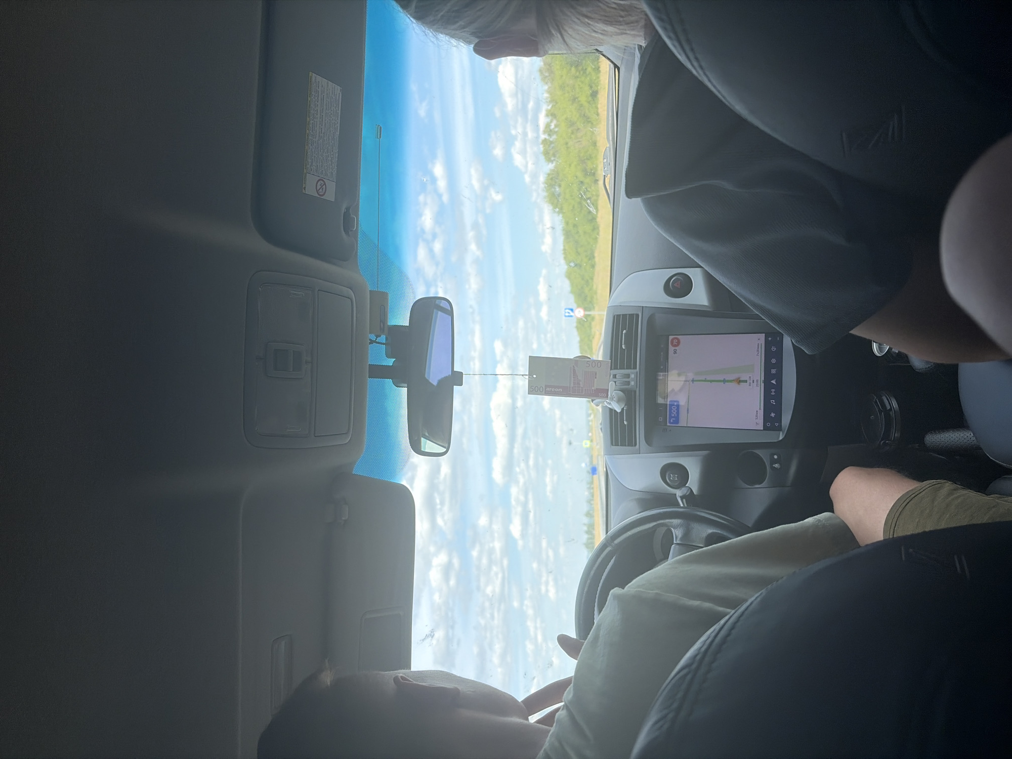 The image shows the interior of a car, viewed from the back seat, looking forward. Two individuals are present; the driver, a person with short hair wearing a light-colored t-shirt and khaki shorts, is visible from the shoulders up. To the right, another person, whose head and shoulder are visible, is wearing a dark t-shirt. The car's dashboard features a navigation screen displaying a map with route information and speed limits of 90 and 70. A car air freshener with a holographic design hangs from the rearview mirror. Outside the car, a bright, cloudy sky is visible above a treeline. The scene appears to be on a road, likely a highway, with a speed limit sign and a parking sign visible in the distance. The overall impression is of a journey in progress on a sunny day. Based on the provided crops and typical car interior details, the location is not definitively Novoe Teneevo, Russia.