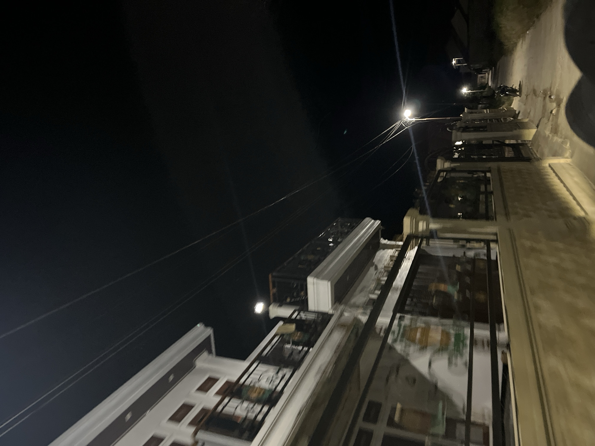 The image depicts a street scene at night in Menanggini, Indonesia. The perspective is tilted, giving a dynamic and somewhat disorienting view of the surroundings. The primary subjects are buildings and the street itself, illuminated by streetlights.
On the right side of the image, a residential building with a light-colored facade and dark-trimmed windows is visible. The building appears to be multi-story, with balconies or architectural elements suggesting balconies. Further along the street, other buildings and structures are visible, some with illuminated windows, indicating indoor activity.
A paved street runs through the scene, appearing wet in some areas, possibly from recent rain. A motorcycle is parked on the side of the street, suggesting local transportation. Overhead, power lines stretch across the dark sky, a common sight in urban or semi-urban areas. The sky is dark, indicating nighttime, and there are no apparent weather phenomena like rain or fog, although the wet street might suggest it.
The overall mood is quiet and subdued, typical of a residential area at night. The artificial lighting from the streetlights casts long shadows and highlights the textures of the buildings and the wet pavement. No people are clearly visible in the image, adding to the sense of stillness. The setting appears to be a street in a neighborhood, likely in a town or city. There is no visible text in the image.