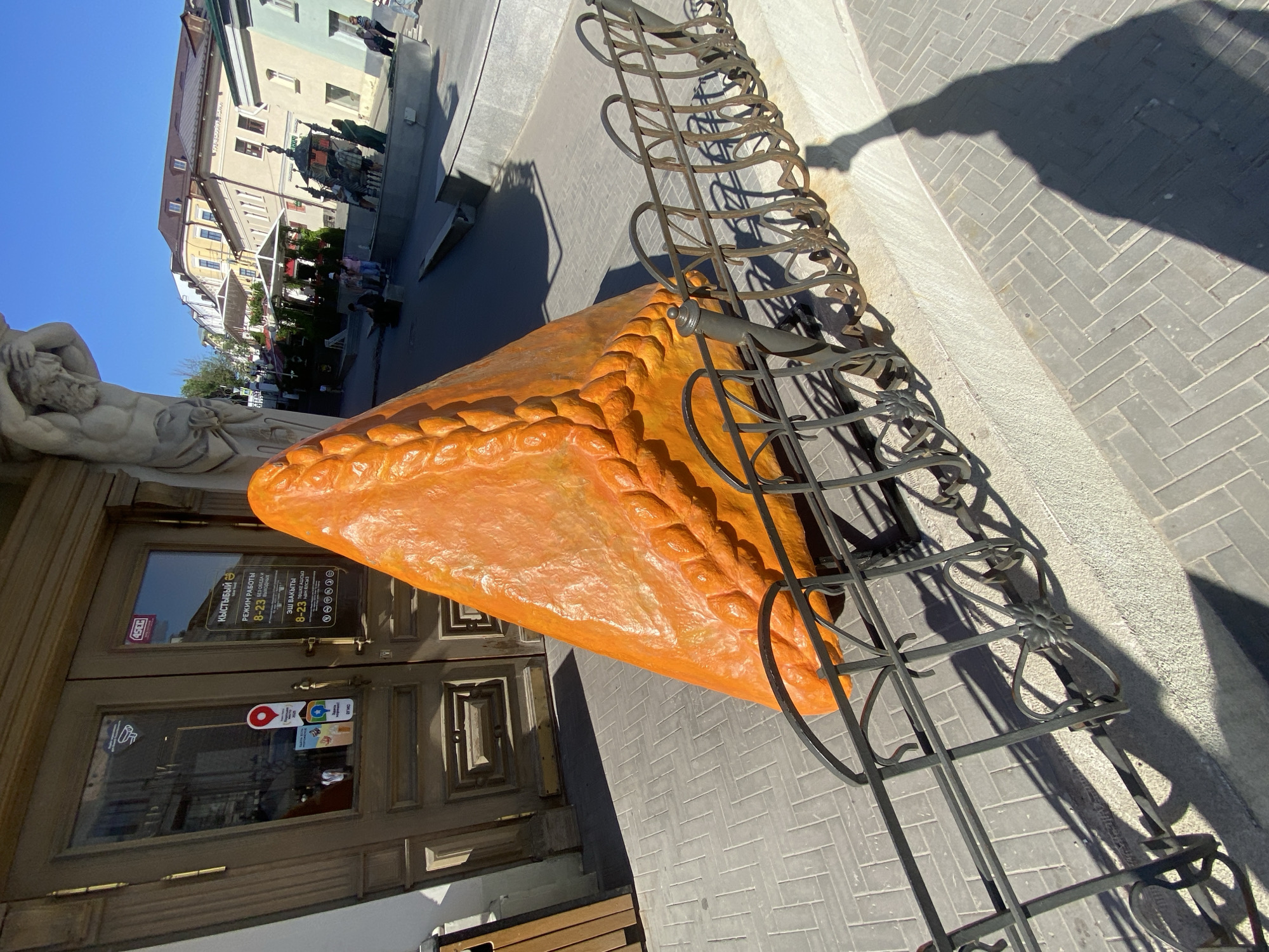 This is an outdoor photograph taken in Kazan, Russia. In the foreground is a large, orange, triangular sculpture resembling a pastry. The sculpture is partially enclosed by a low, decorative black metal fence. To the left of the sculpture is a building with a wooden door. A sign is visible on the door, written in Russian, with operating hours from 8-23. Next to the door is a white statue of a reclining man. In the background, across a paved street, are buildings with shops and restaurants. People are visible on the street. The sky is blue. The image is taken during the day, with strong shadows indicating bright sunlight. The image seems to be rotated to the left.