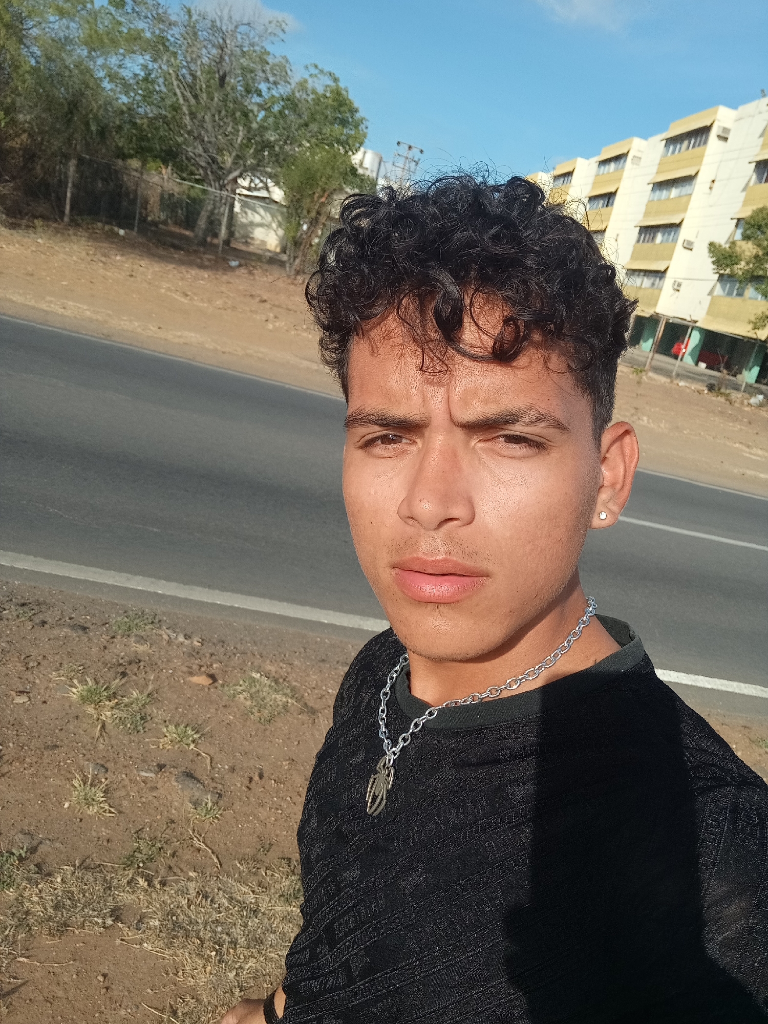 A young man with dark, curly hair poses for a picture outdoors under a clear blue sky. He has a neutral to slightly serious expression and wears a black, textured long-sleeved shirt. A prominent silver chain necklace with a unique, abstract pendant resembling a stylized spider or intertwined design hangs around his neck. A small silver stud earring is visible in his left ear.
He is standing on the dirt shoulder of a paved road, which features a distinct white line. The ground is dry, with sparse patches of light green and brown vegetation. In the background to his left, several trees and a fence are visible. To his right, a large, multi-story building with yellow and white facades and multiple windows, some fitted with air conditioning units, stands under the bright sun. The architecture suggests an older structure, possibly an apartment or commercial building.
The image was taken during the day, likely late morning or early afternoon, given the bright, direct sunlight and pronounced shadows cast on the man's shirt and the ground. The weather appears clear and sunny. The setting suggests an urban or semi-urban environment in Altagracia, Venezuela, characterized by a blend of dry natural landscape and older infrastructure. No visible text is discernible in the image.