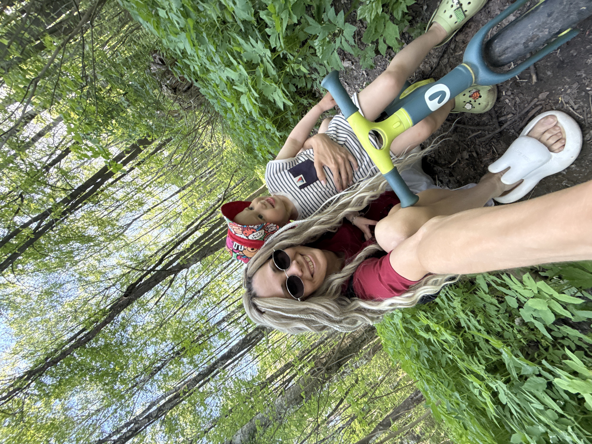 The image appears to be a selfie taken by a woman with a child, possibly in a wooded area. The woman has long blonde hair and is wearing sunglasses and a red top. The child is wearing a striped shirt and a colorful hat. They are both lying on the ground amidst greenery, and a yellow and teal-colored child's scooter is partially visible. The environment suggests a forest or park-like setting, with trees and lush foliage surrounding them. No further location-specific details are discernible from the image.