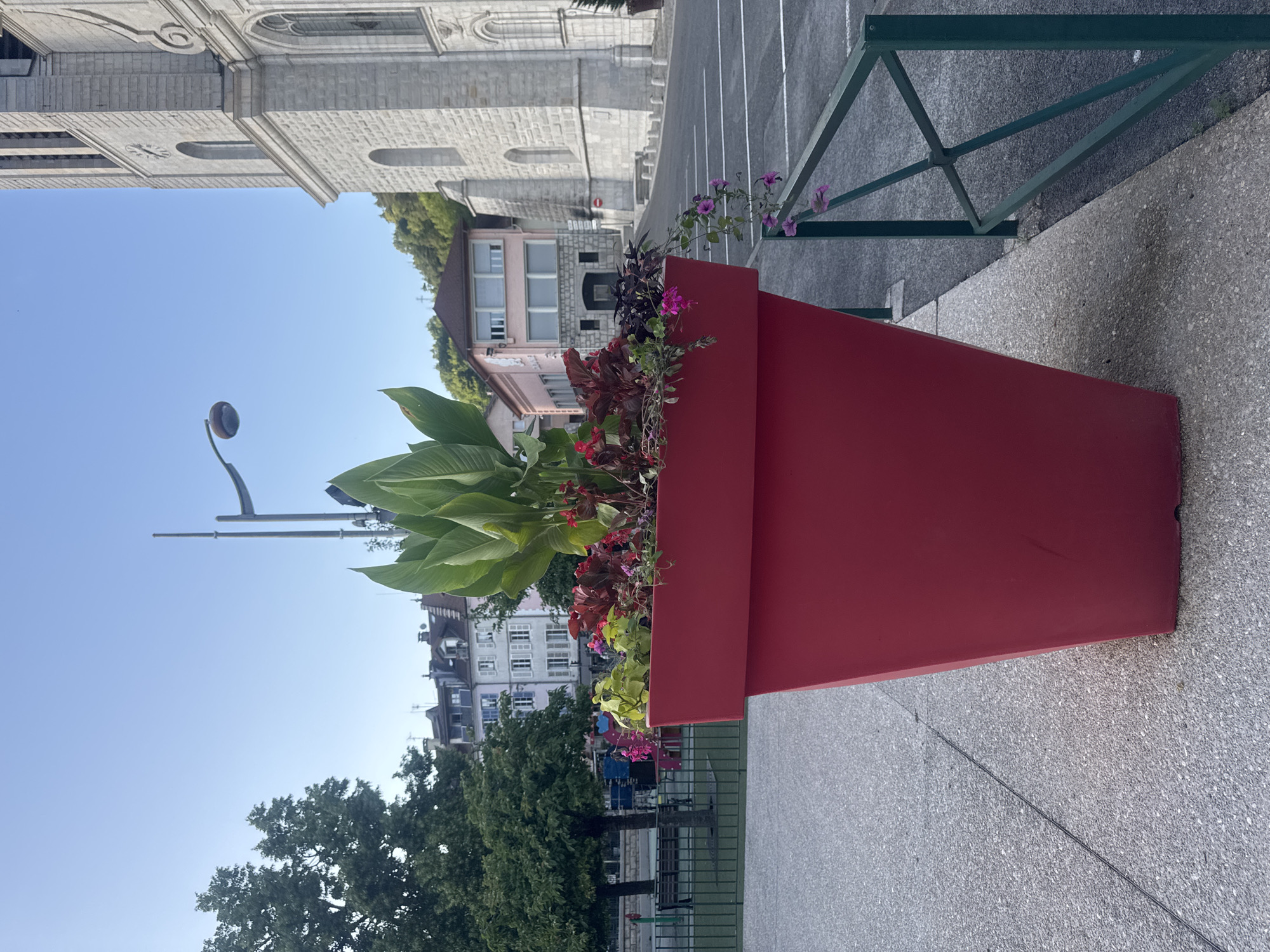 This is a medium-shot photograph of a large, bright red, trapezoidal planter filled with a variety of plants, including large green leaves and smaller flowering plants, situated on a paved sidewalk. A dark green metal railing with a crisscross pattern runs horizontally across the lower right portion of the frame, with a single pink flower hanging over it. Behind the planter, a street is visible with white parking lines. In the background, a large, light-colored stone building, likely a church due to its architectural style with arched windows and a bell tower, dominates the right side of the image. To the left of the church and behind the planter, there are several multi-story buildings with varying architectural styles and colors. A large tree with green foliage is visible on the far left, and a lamppost with a distinctive curved arm and a rounded light fixture is present in the upper center. The sky is clear and blue. The scene appears to be in an urban or town setting, possibly a public square or street in Saint-Claude, France, given the architecture. No people are visible in the frame.